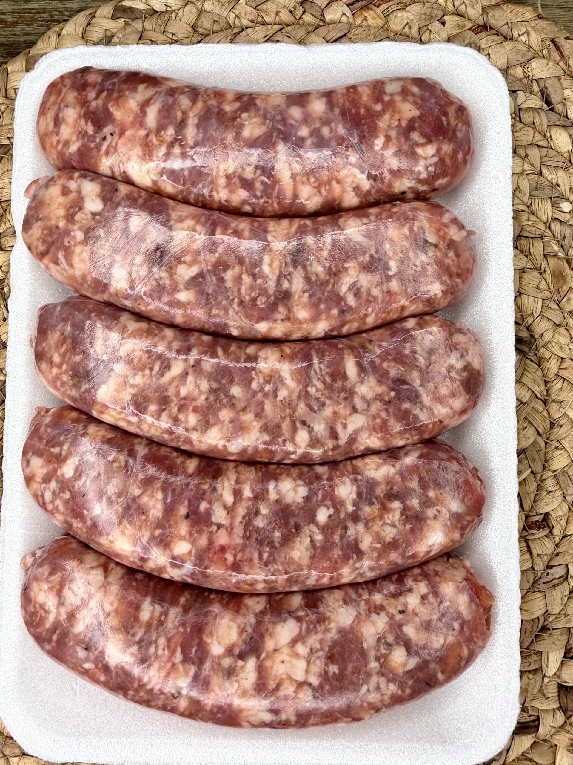Five raw sausages in clear casings are arranged in a row on a white foam tray, placed on a woven mat background.