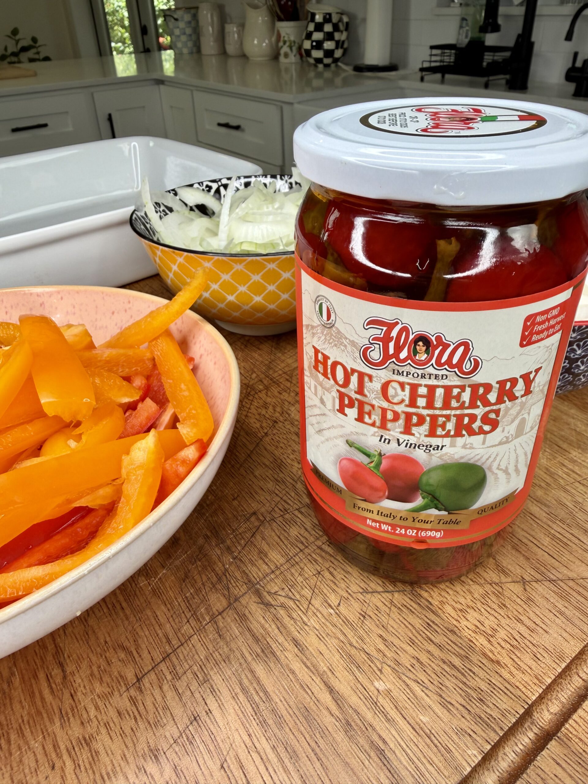A jar of Flora Hot Cherry Peppers in vinegar sits on a wooden counter next to a bowl of sliced orange bell peppers and a bowl of sliced onions in a kitchen setting.