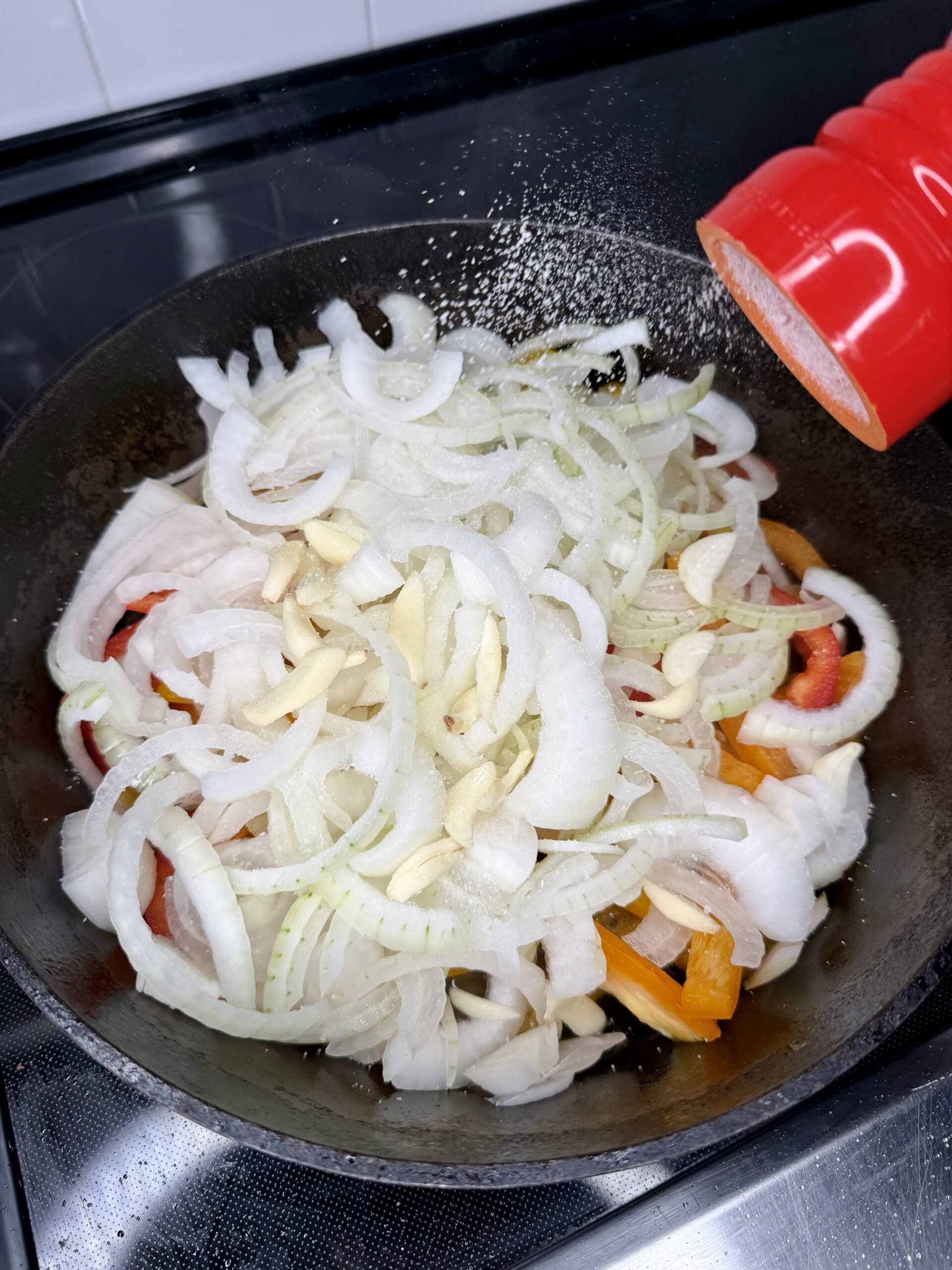 Sliced onions, garlic, and bell peppers cooking in a black pan on a stovetop, with salt being sprinkled from a red shaker.