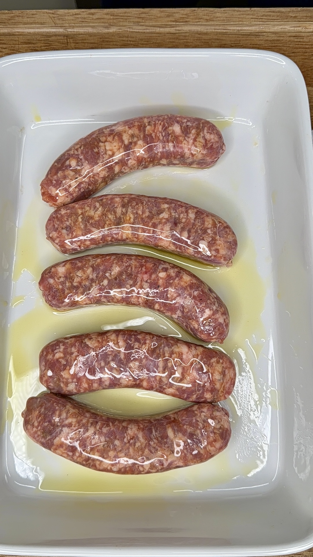 Five uncooked sausages coated in oil are arranged in a white rectangular baking dish on a wooden surface.