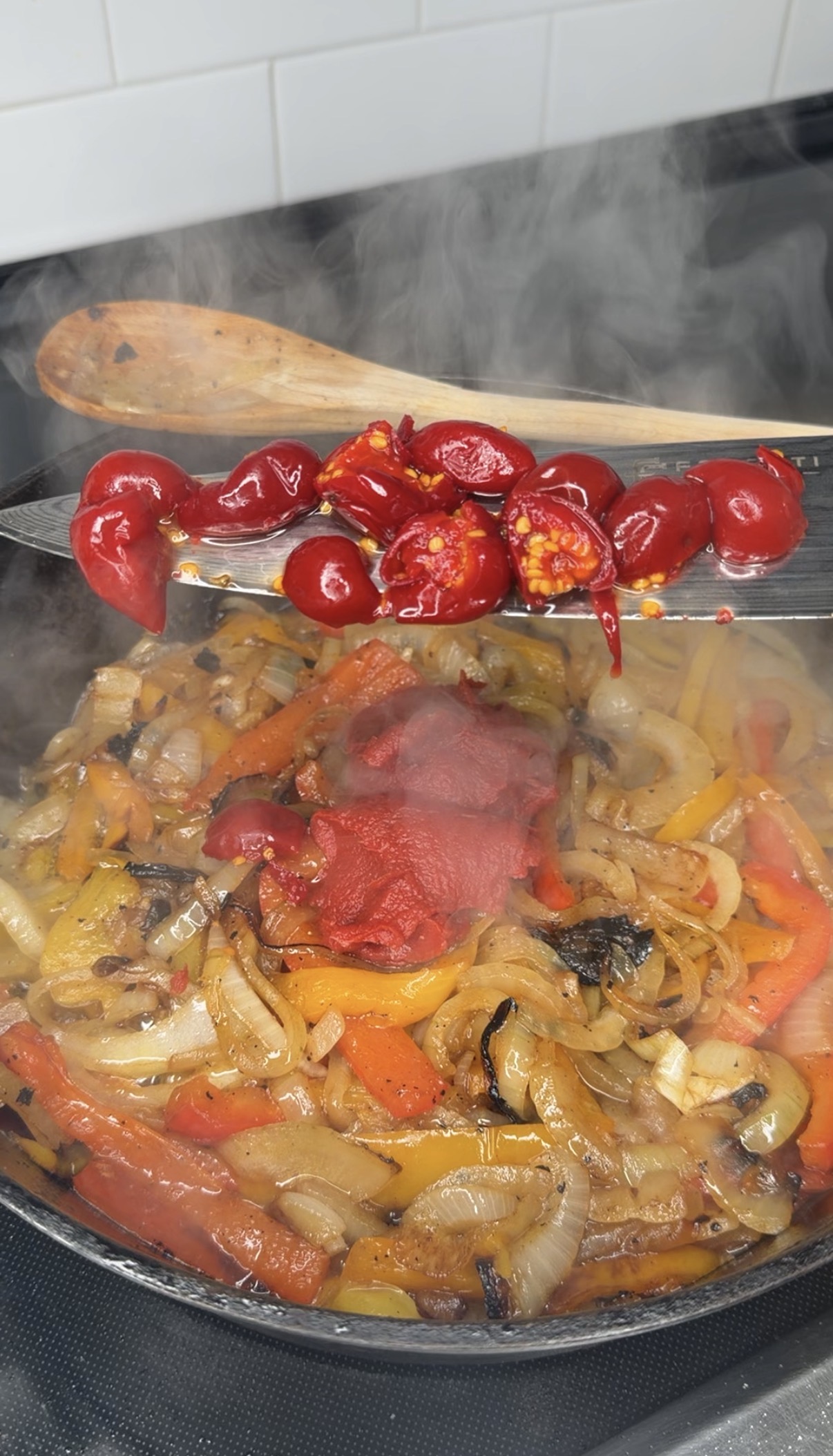A close-up of sautéed onions, bell peppers, and mushrooms in a pan with a spoonful of red cherry peppers and tomato paste being added, releasing steam, with a wooden spoon resting nearby.