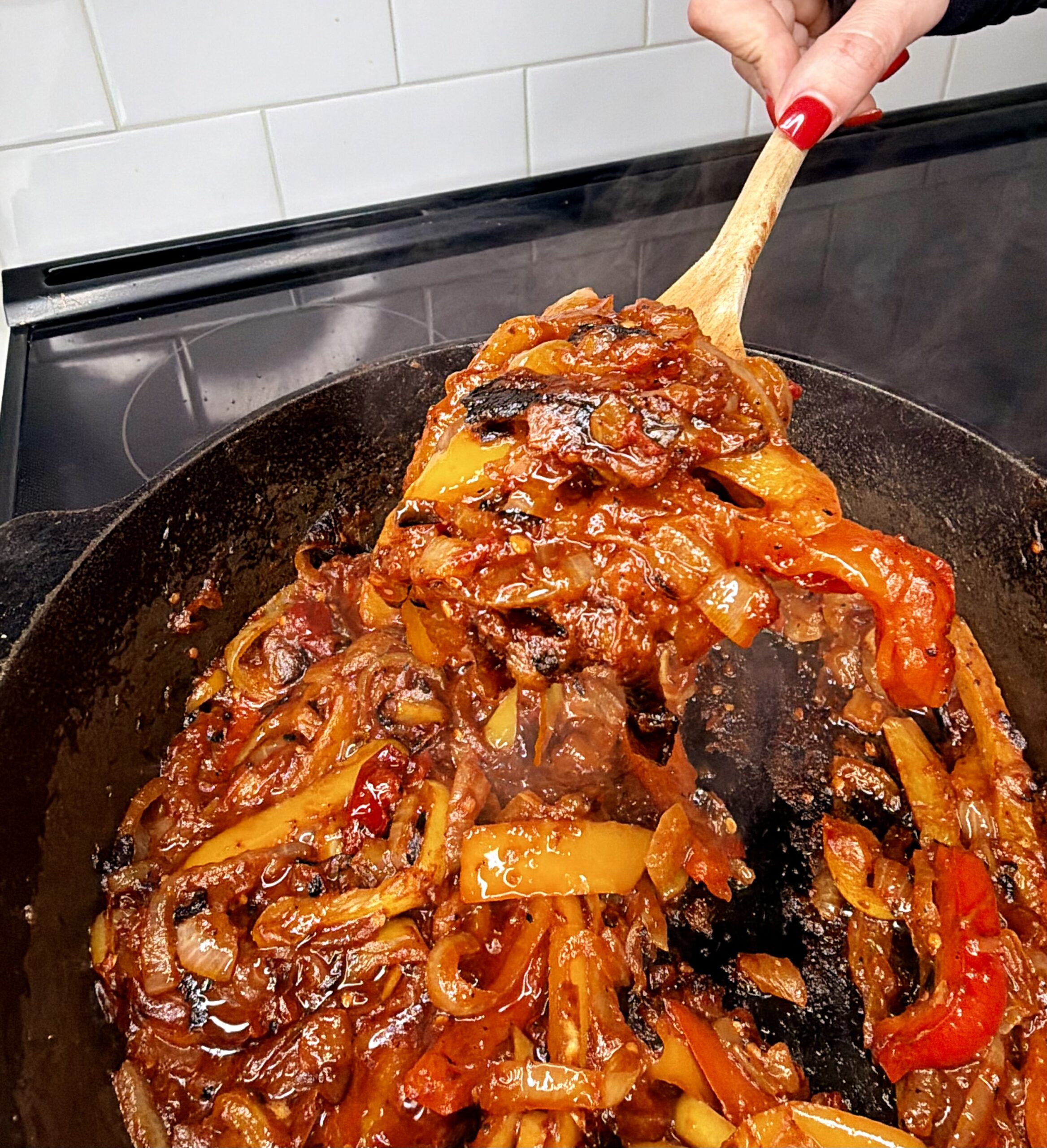 A hand holding a wooden spoon stirs a cast iron skillet filled with sautéed onions, red and yellow bell peppers, and tomato sauce on a stovetop. Steam rises from the hot, colorful mixture.