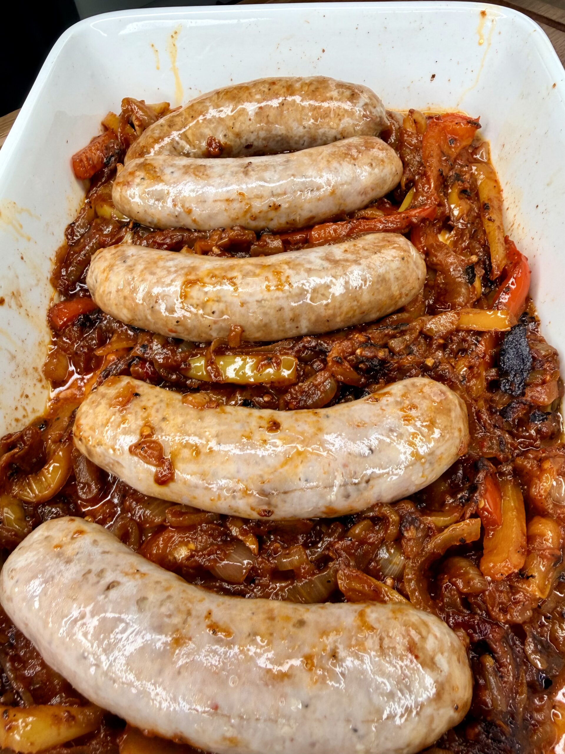 Five uncooked sausages arranged in a white baking dish on top of a bed of sautéed peppers, onions, and tomato sauce, ready to be baked.