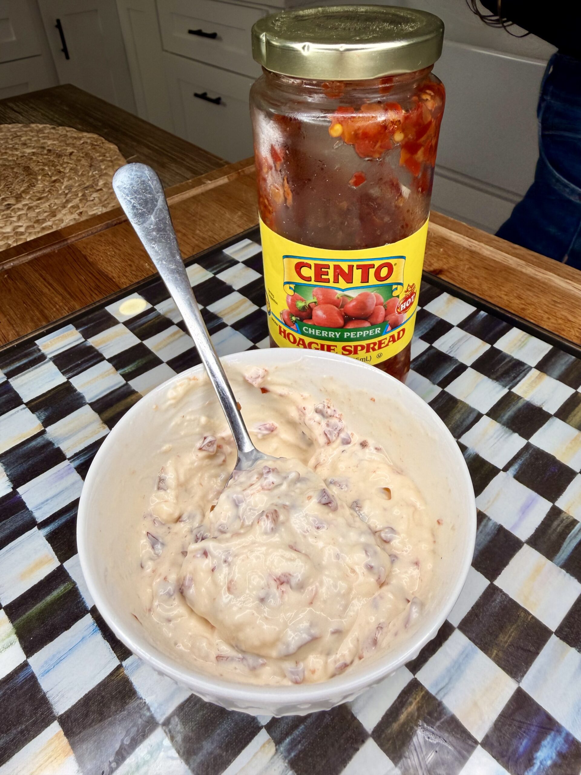 A bowl of creamy dip with a spoon rests on a checkered surface, next to an open jar of Cento Cherry Pepper Hoagie Spread.