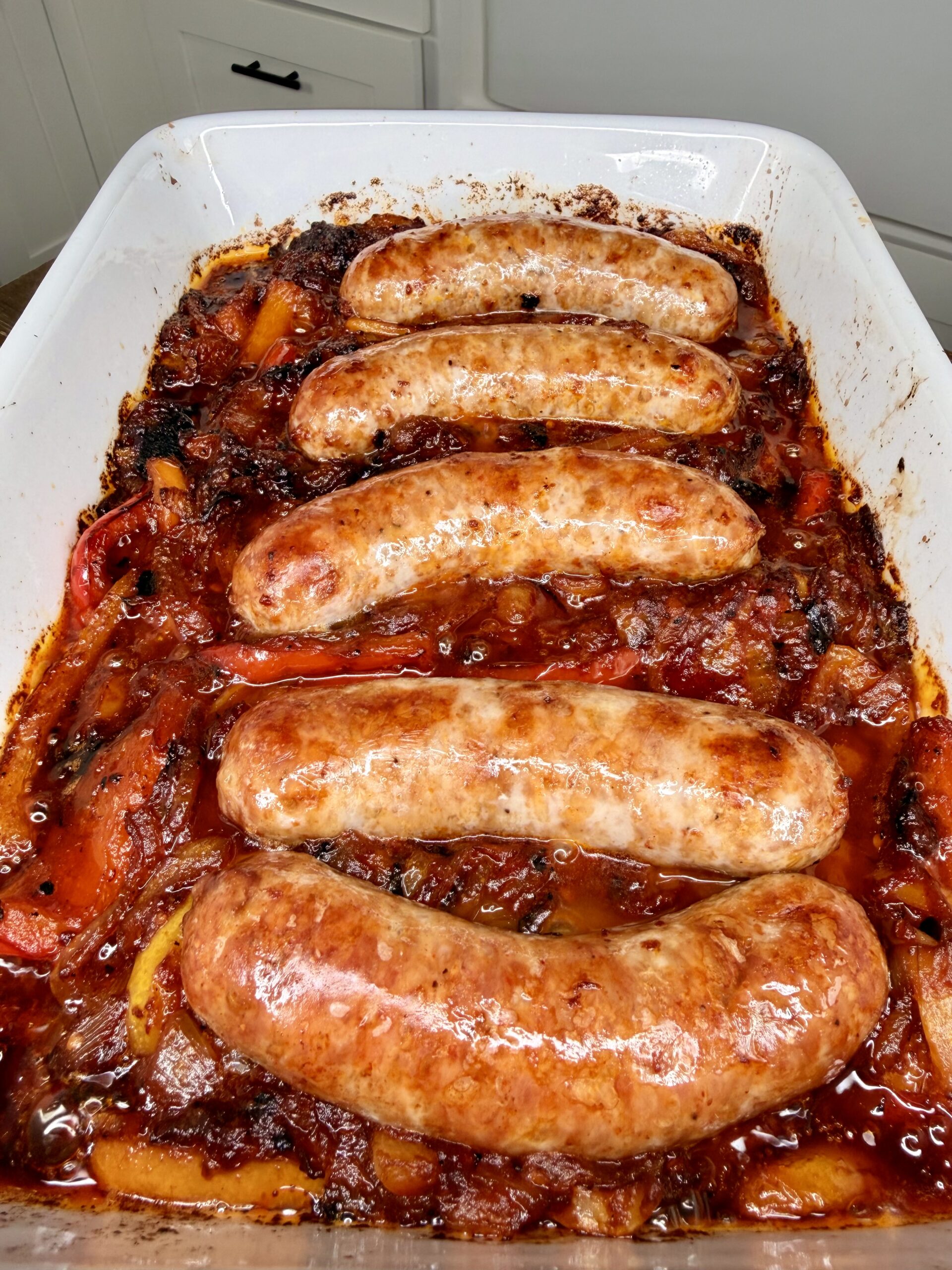 Five cooked sausages on top of roasted bell peppers and onions in a white baking dish, surrounded by a rich, tomato-based sauce.