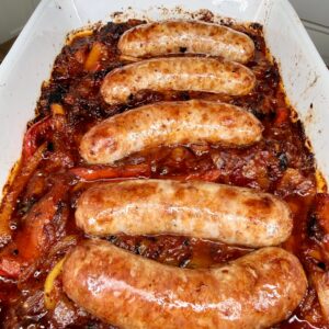 Five browned sausages baked on top of a bed of roasted bell peppers, onions, and tomato sauce in a white casserole dish. The dish looks juicy and caramelized with crispy edges.