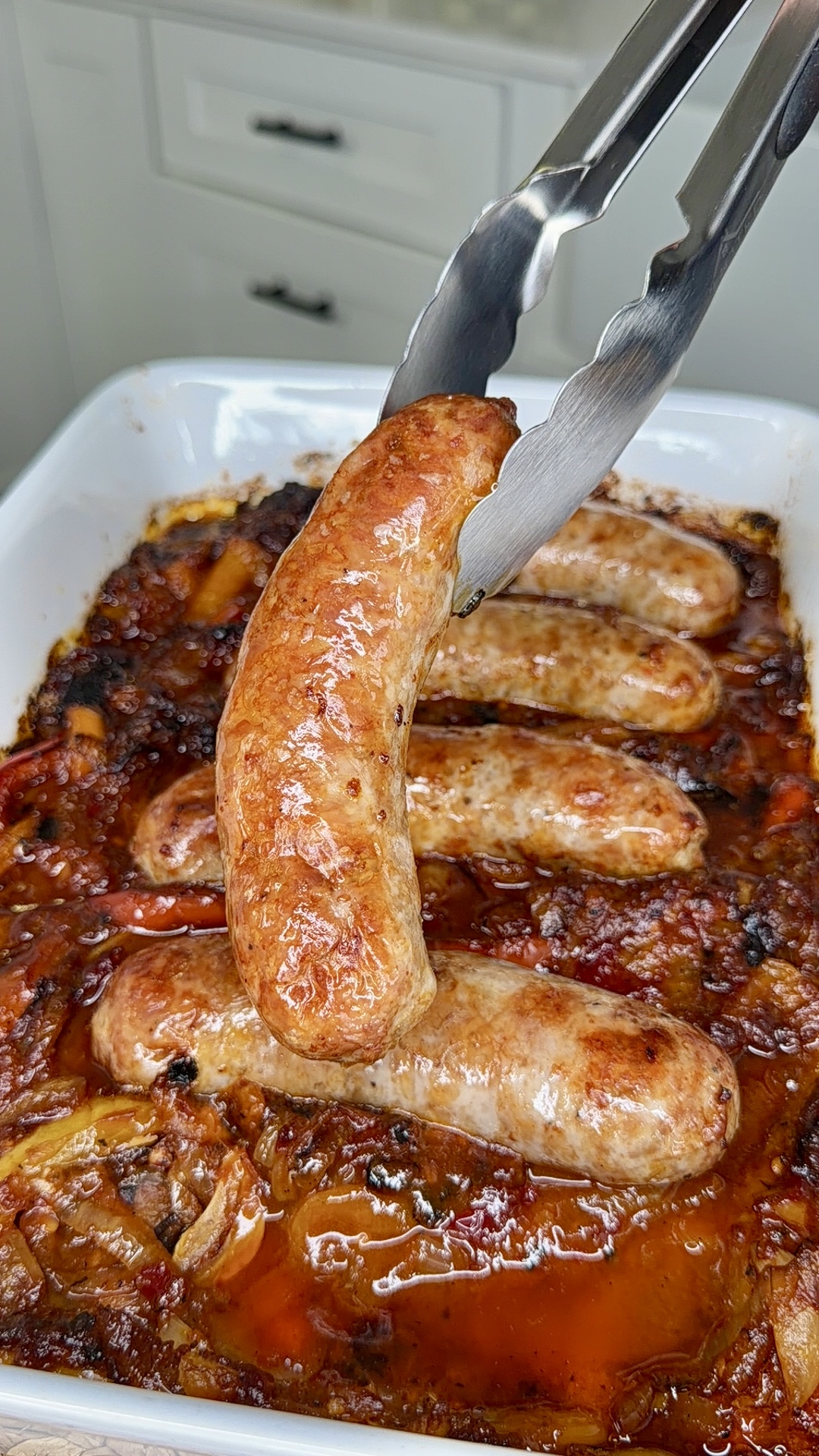 A close-up of metal tongs lifting a cooked sausage from a white baking dish filled with sausages and a mixture of roasted vegetables and sauce. The sausages are browned and the dish appears freshly baked.
