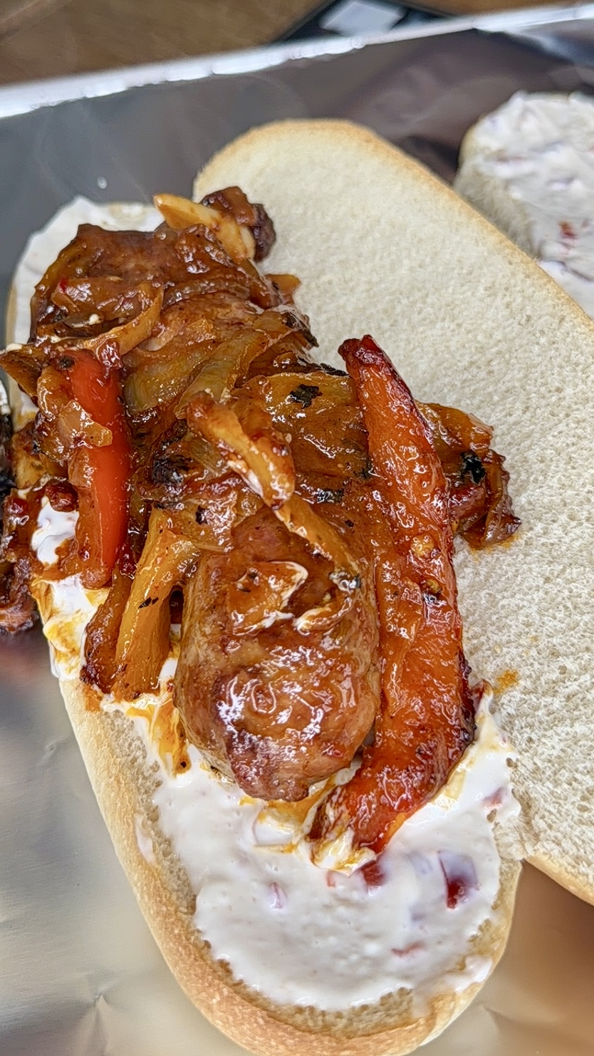 A sausage in a bun topped with sautéed onions and red bell peppers, with a generous spread of creamy white sauce on the bun.