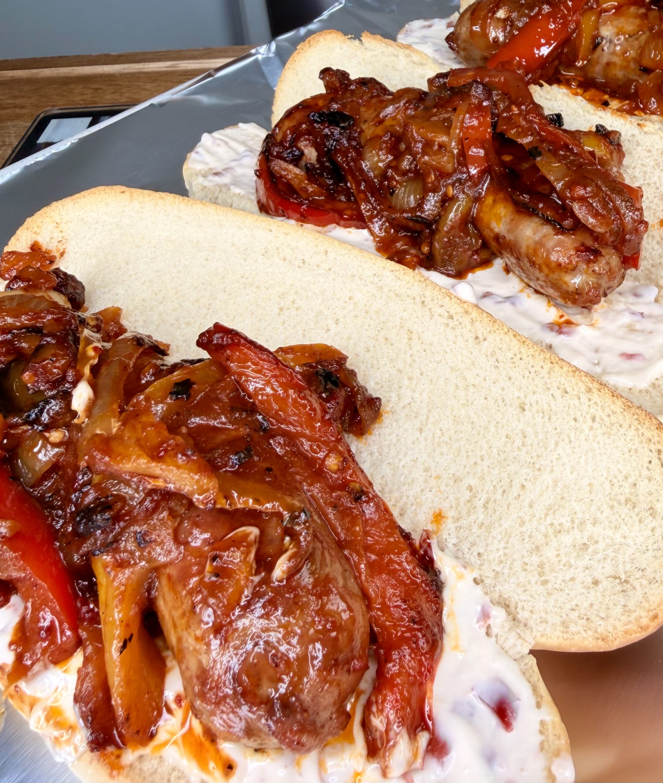 Two hoagie rolls filled with sausages, sautéed red peppers, and onions, topped with a creamy white sauce, served on a metal tray.