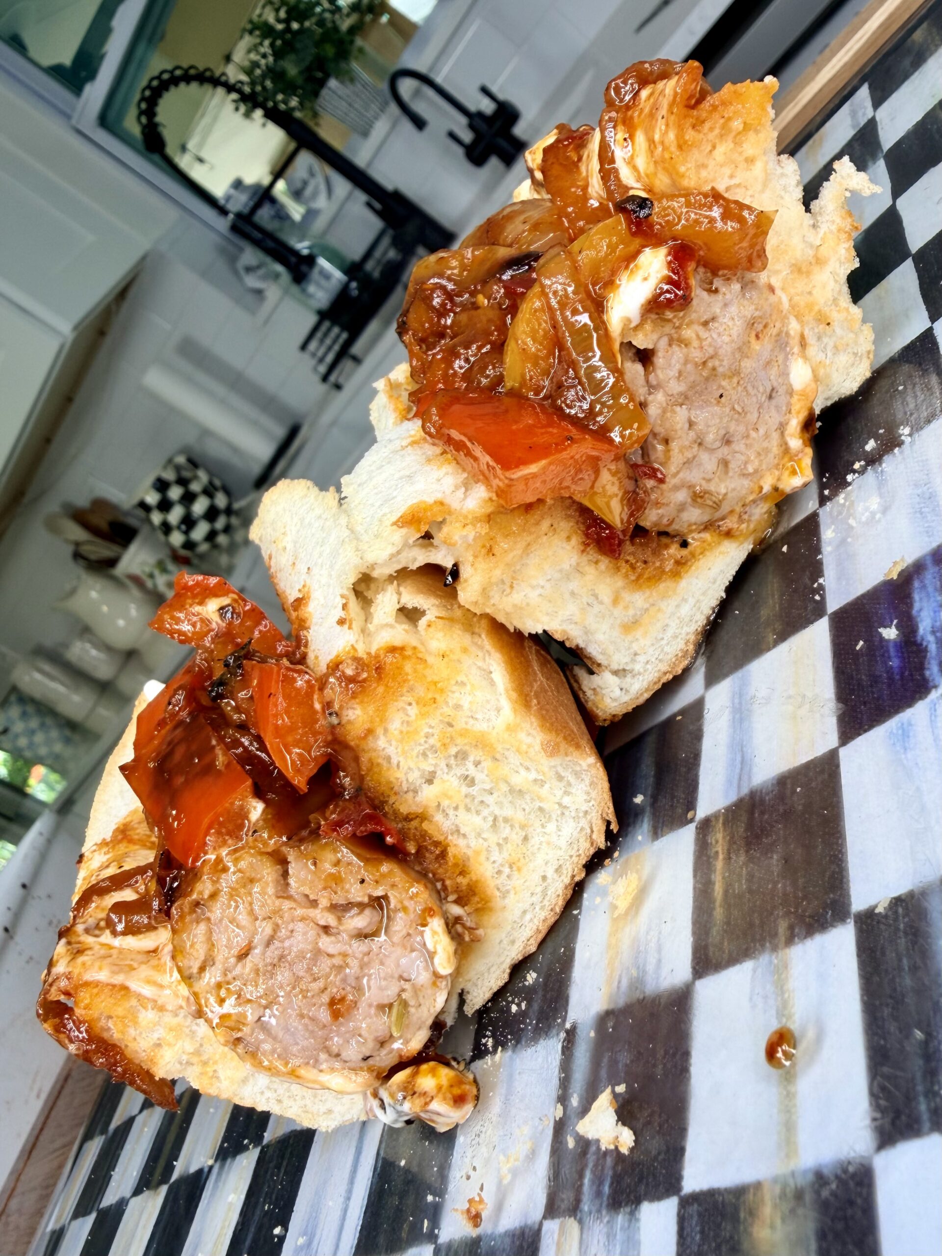 A close-up of a sliced meatball sandwich on crusty bread, topped with sautéed onions and red peppers, set on a black-and-white checkered cutting board in a bright kitchen.