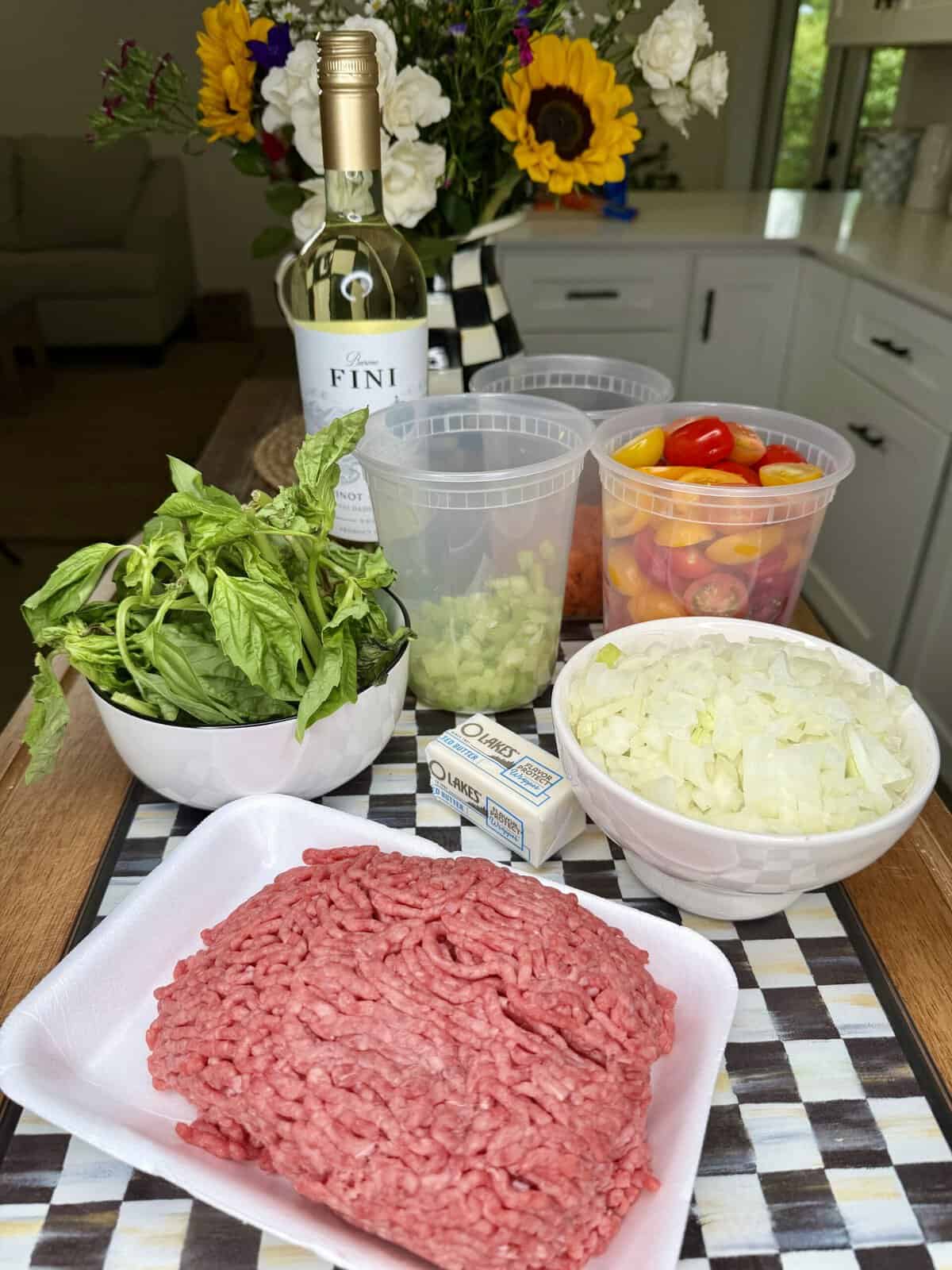 A tray of ground beef, chopped onions, celery, colorful cherry tomatoes, fresh basil, butter, and a bottle of white wine are arranged on a kitchen counter with flowers in the background.