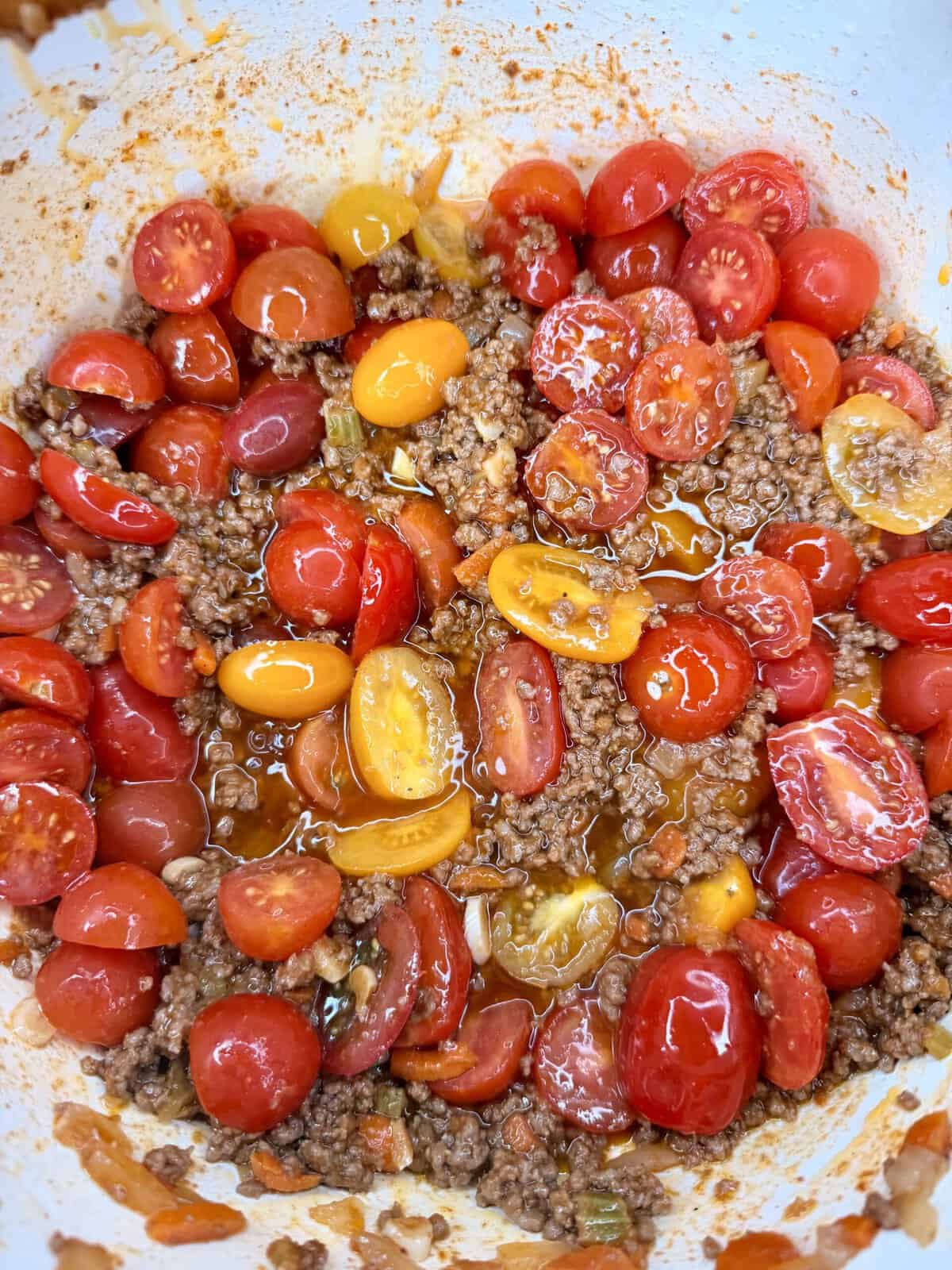 A close-up of halved red and yellow cherry tomatoes mixed with cooked ground beef and juices in a white bowl.