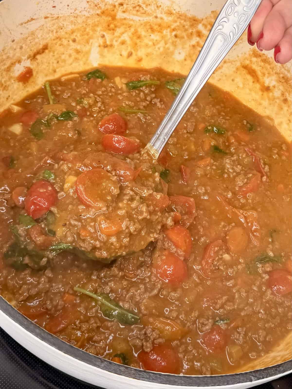 A close-up of a pot of hearty tomato-based meat sauce with ground beef, cherry tomatoes, and spinach, being stirred with a silver ladle by a person with red-painted nails.