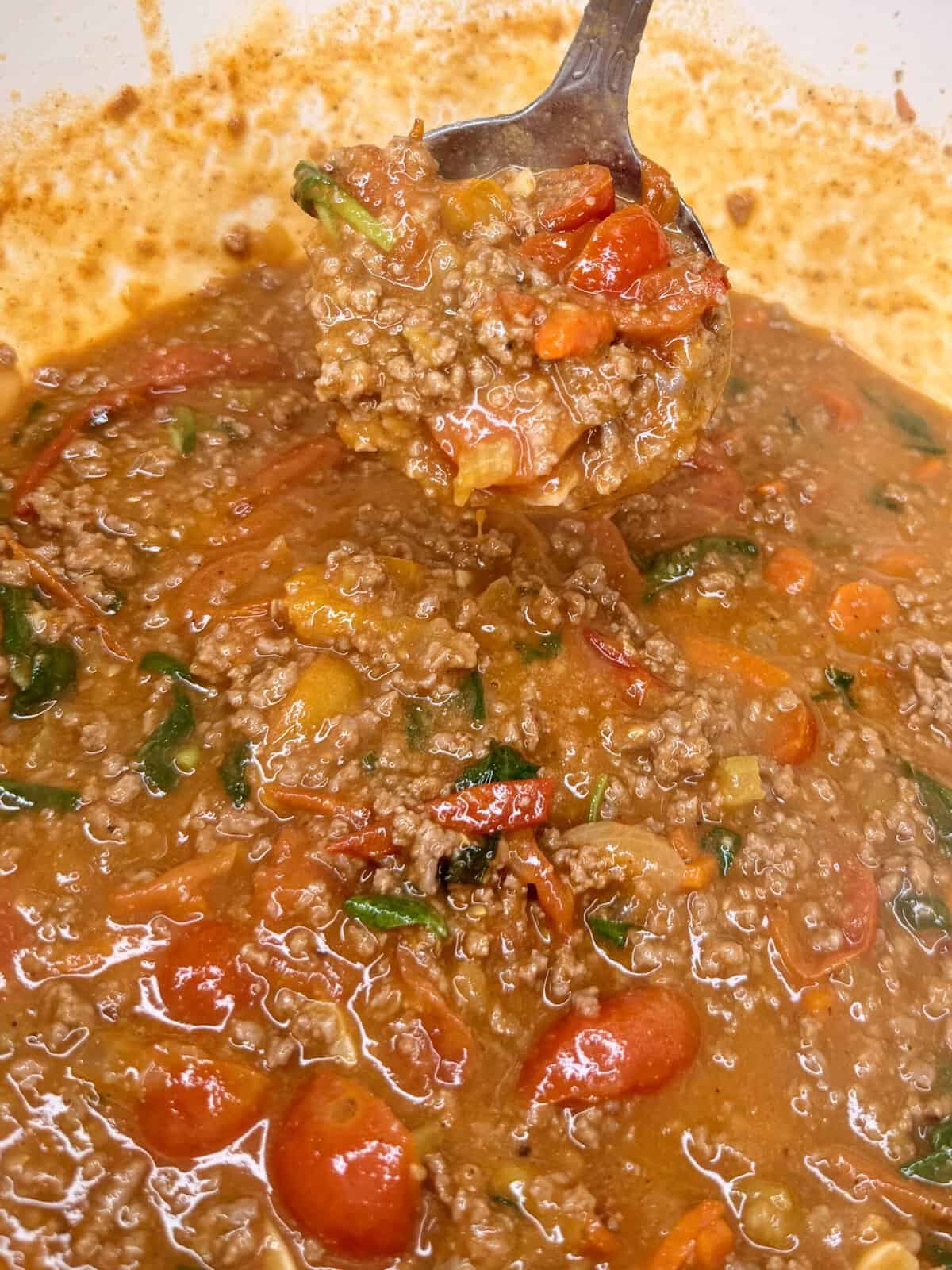 A close-up of a spoonful of hearty chili with ground beef, halved cherry tomatoes, spinach, and a rich, reddish-brown sauce in a pot.