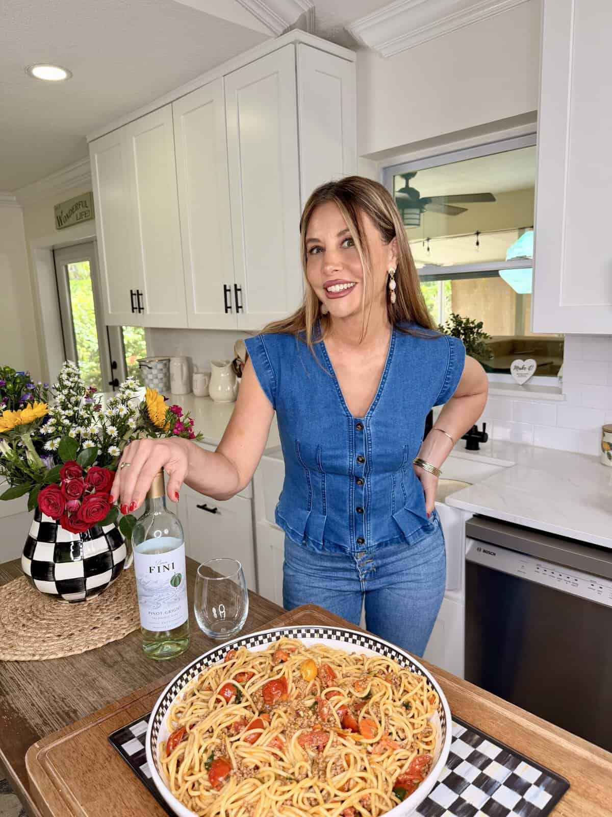 A woman in a blue denim top stands in a bright kitchen, smiling and holding a bottle of white wine next to a glass. In front of her is a large plate of pasta with tomatoes. A bouquet of flowers sits on the counter.