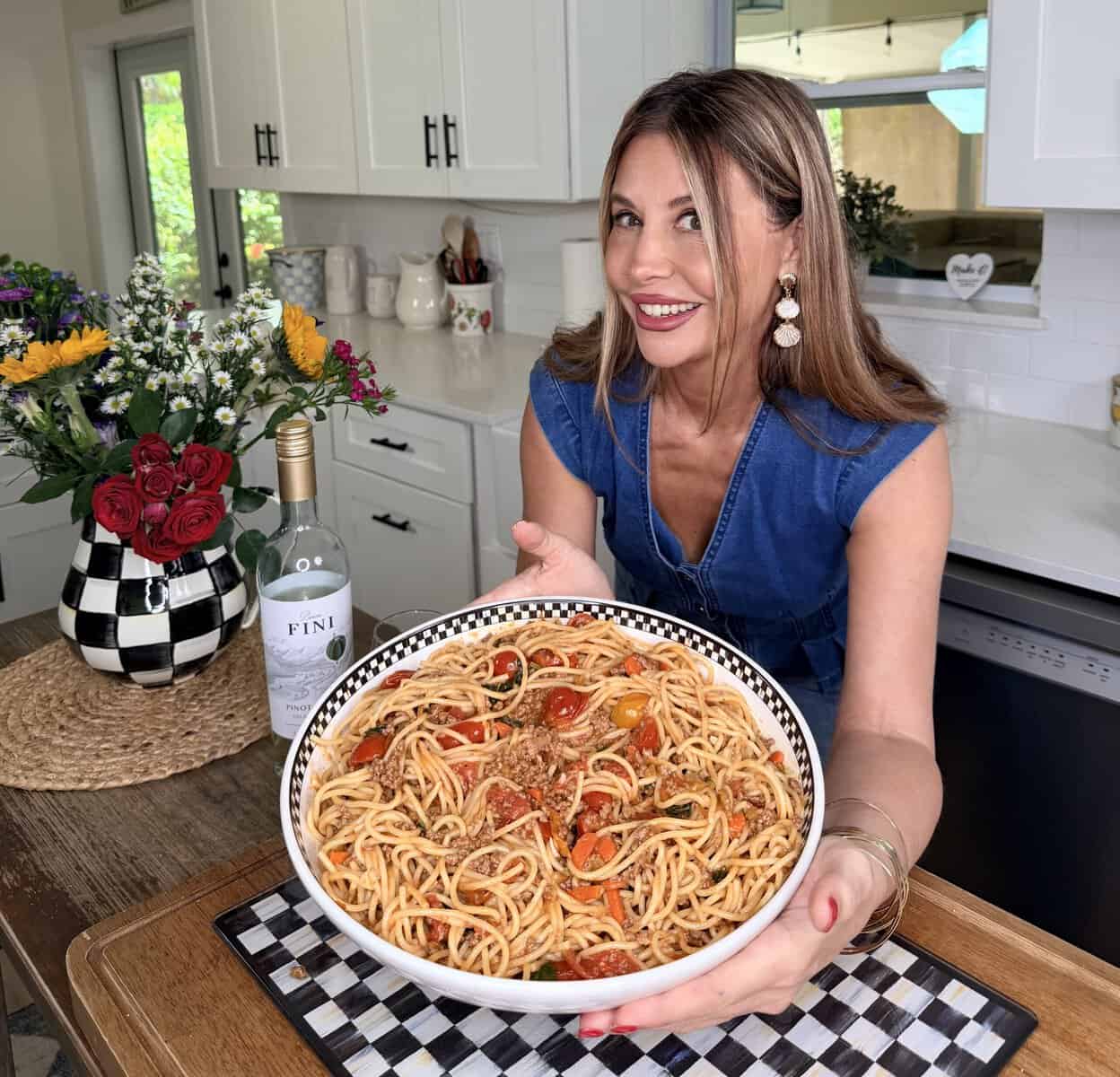 A smiling woman in a blue dress holds a large bowl of spaghetti with tomatoes and herbs in a bright kitchen. A bottle of wine and a vase of flowers are on the counter beside her.