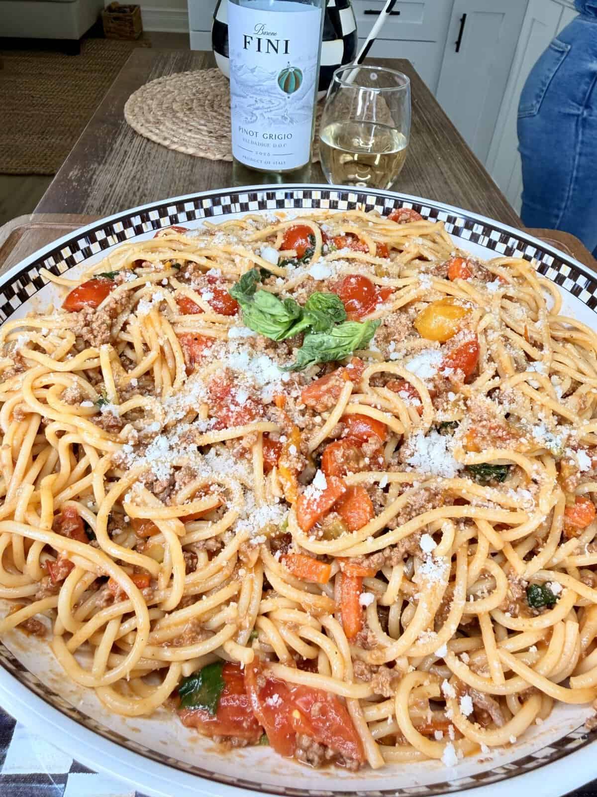 A large plate of spaghetti with meat sauce, cherry tomatoes, basil, and grated cheese sits on a table next to a glass of white wine and a bottle of Fini Pinot Grigio.