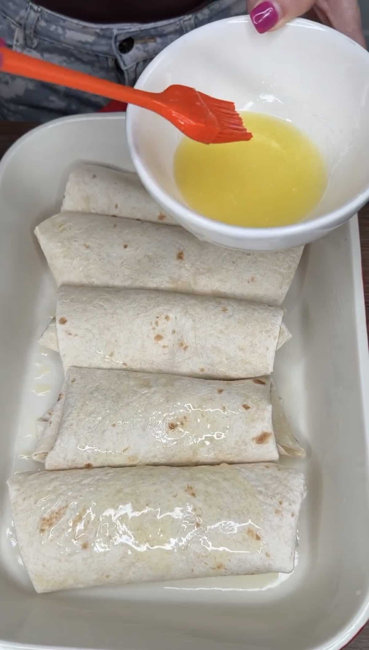 Four rolled tortillas are arranged in a white baking dish. A hand is brushing melted butter from a small white bowl onto the tortillas.