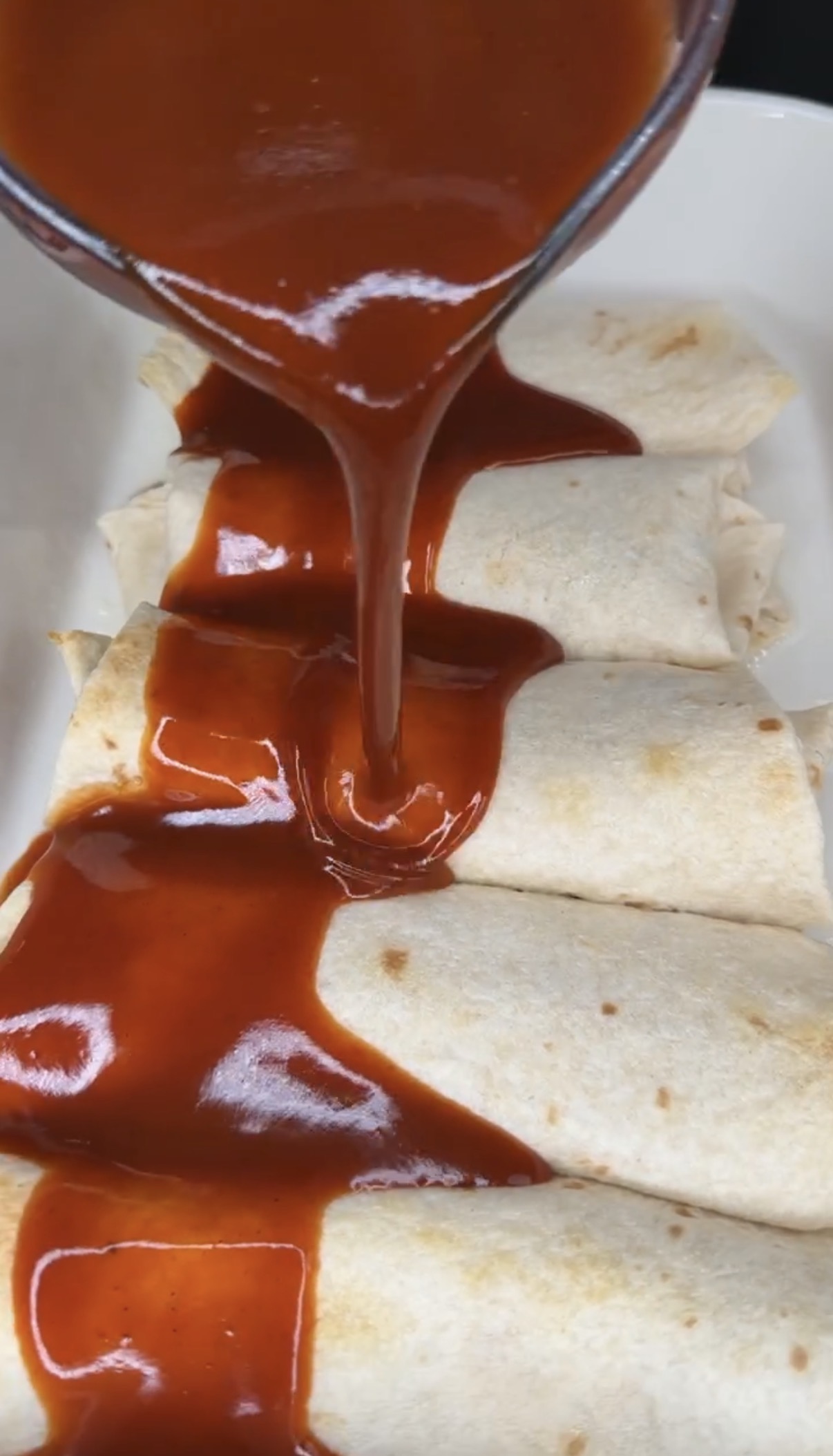 A close-up of enchiladas in a white dish as rich red enchilada sauce is being poured over the top of the rolled tortillas.