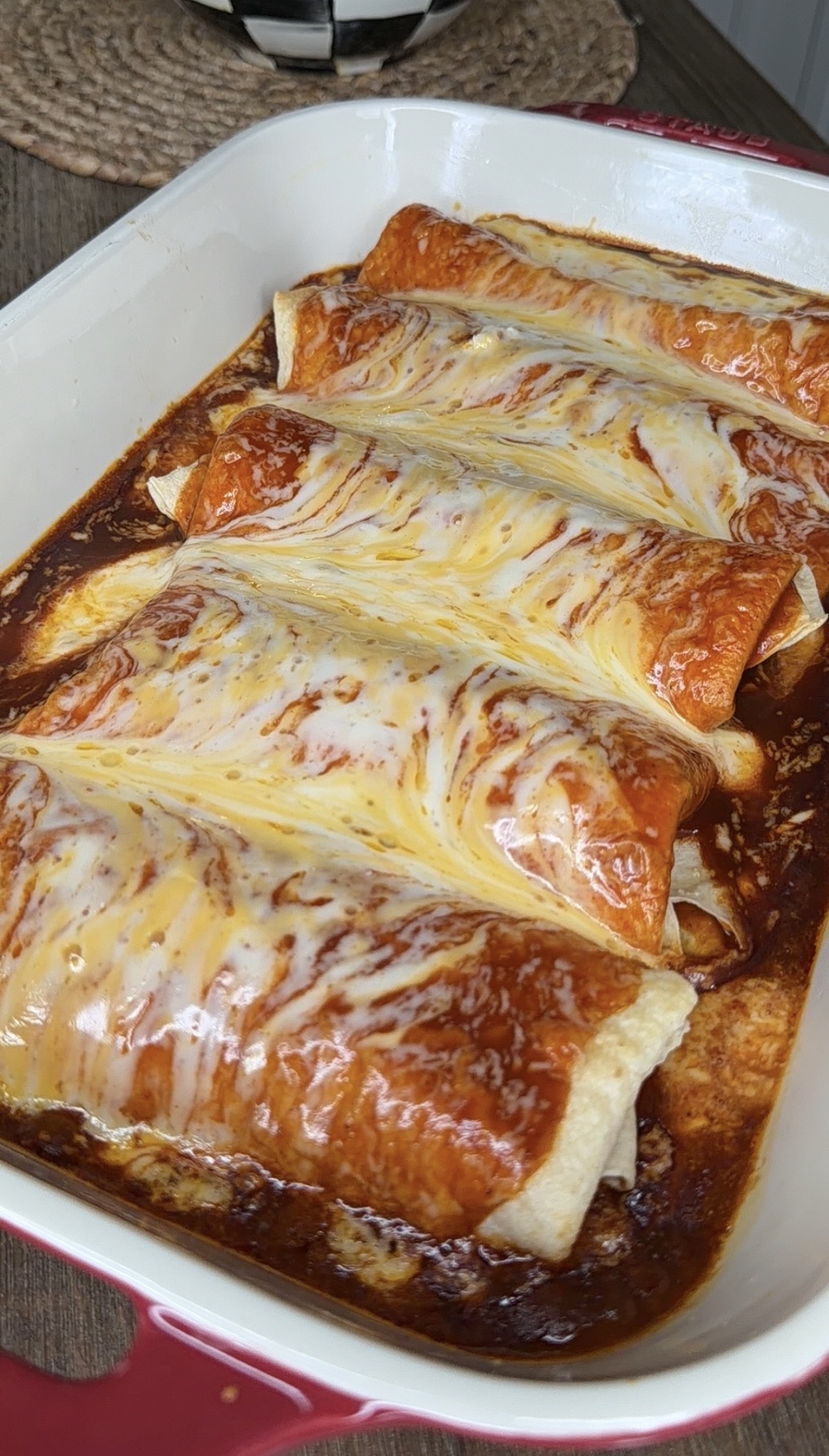 A baking dish filled with five enchiladas covered in melted cheese and red sauce, fresh out of the oven and placed on a wooden table.
