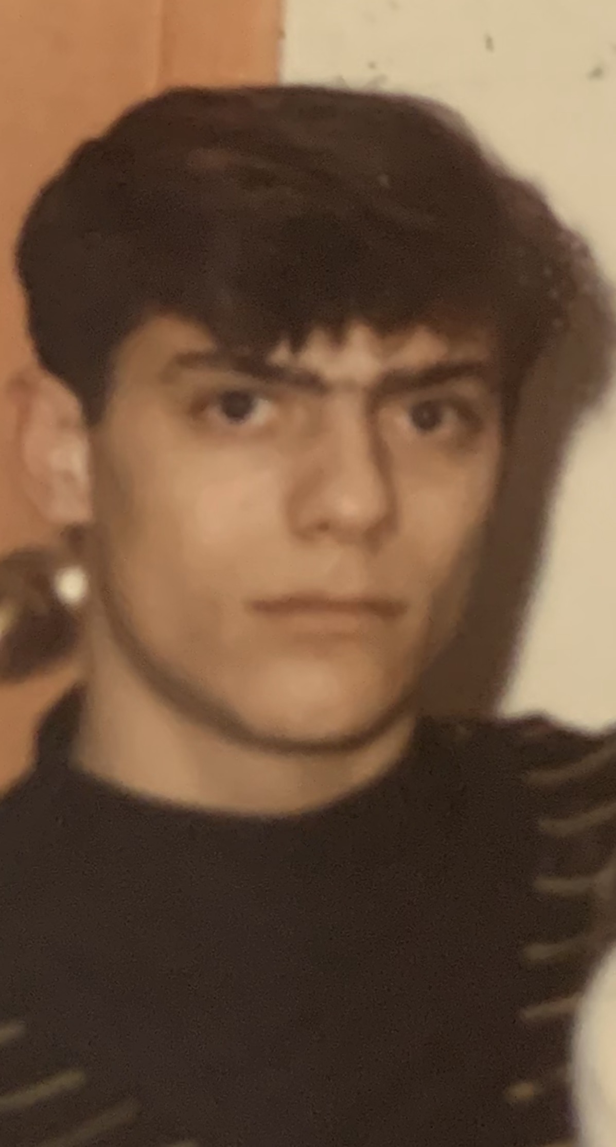 A young person with short dark hair and a serious expression looks at the camera. They are wearing a dark top, and the softly blurred background features light brown tones, reminiscent of the earthy hues in Sicilian Potato Salad.