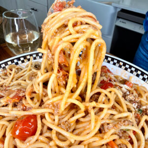 A close-up of a fork lifting Summer Spaghetti Bolognese with tomato and meat sauce from a patterned plate, with a glass of white wine and a kitchen in the background.