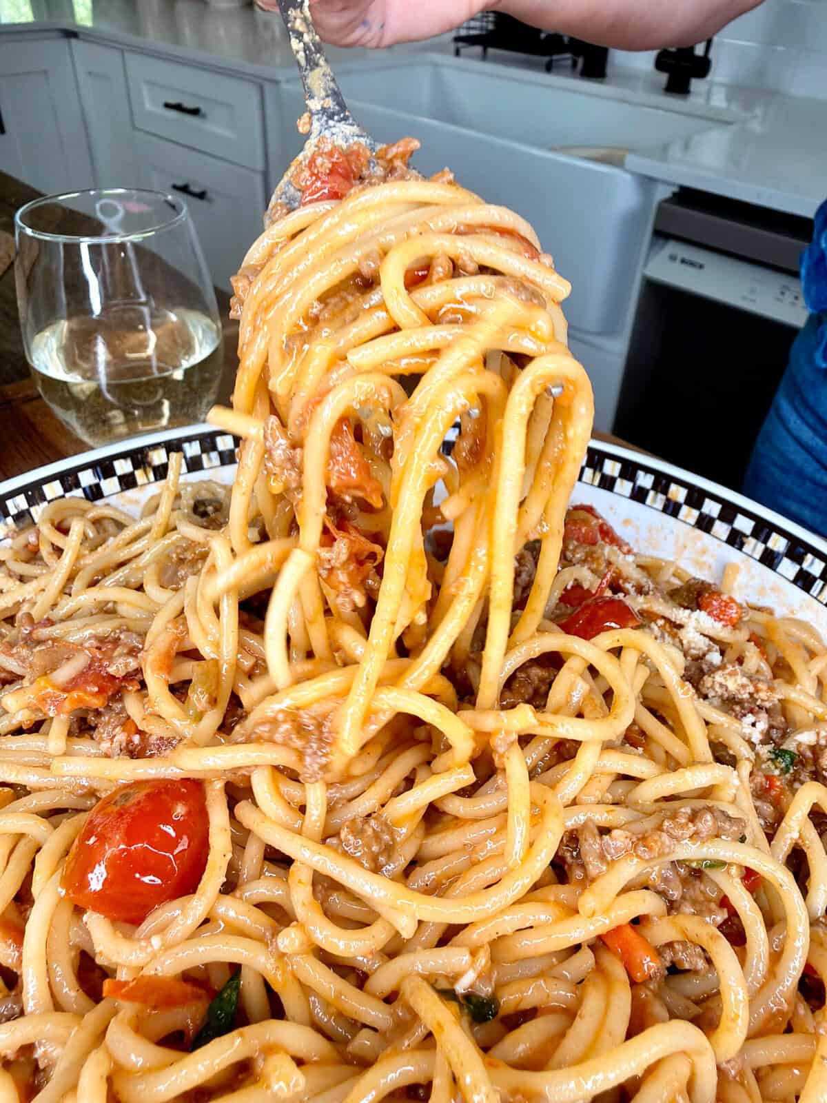 A close-up of a fork twirling spaghetti with tomato sauce, ground meat, and cherry tomatoes over a large plate. A glass of white wine sits in the background on a kitchen counter.