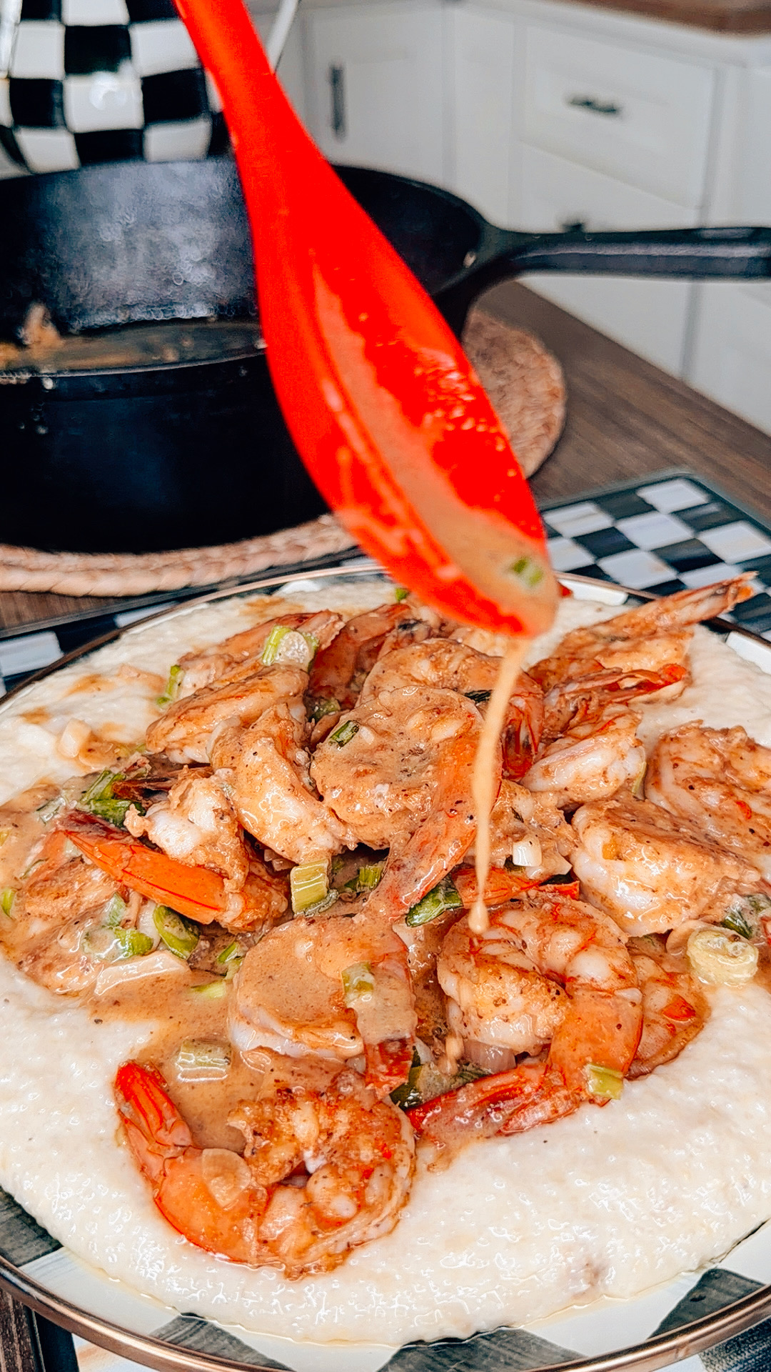 A red spoon drizzles creamy sauce over a plate of shrimp and grits, garnished with chopped green onions, with a cast iron pan and kitchen in the background.