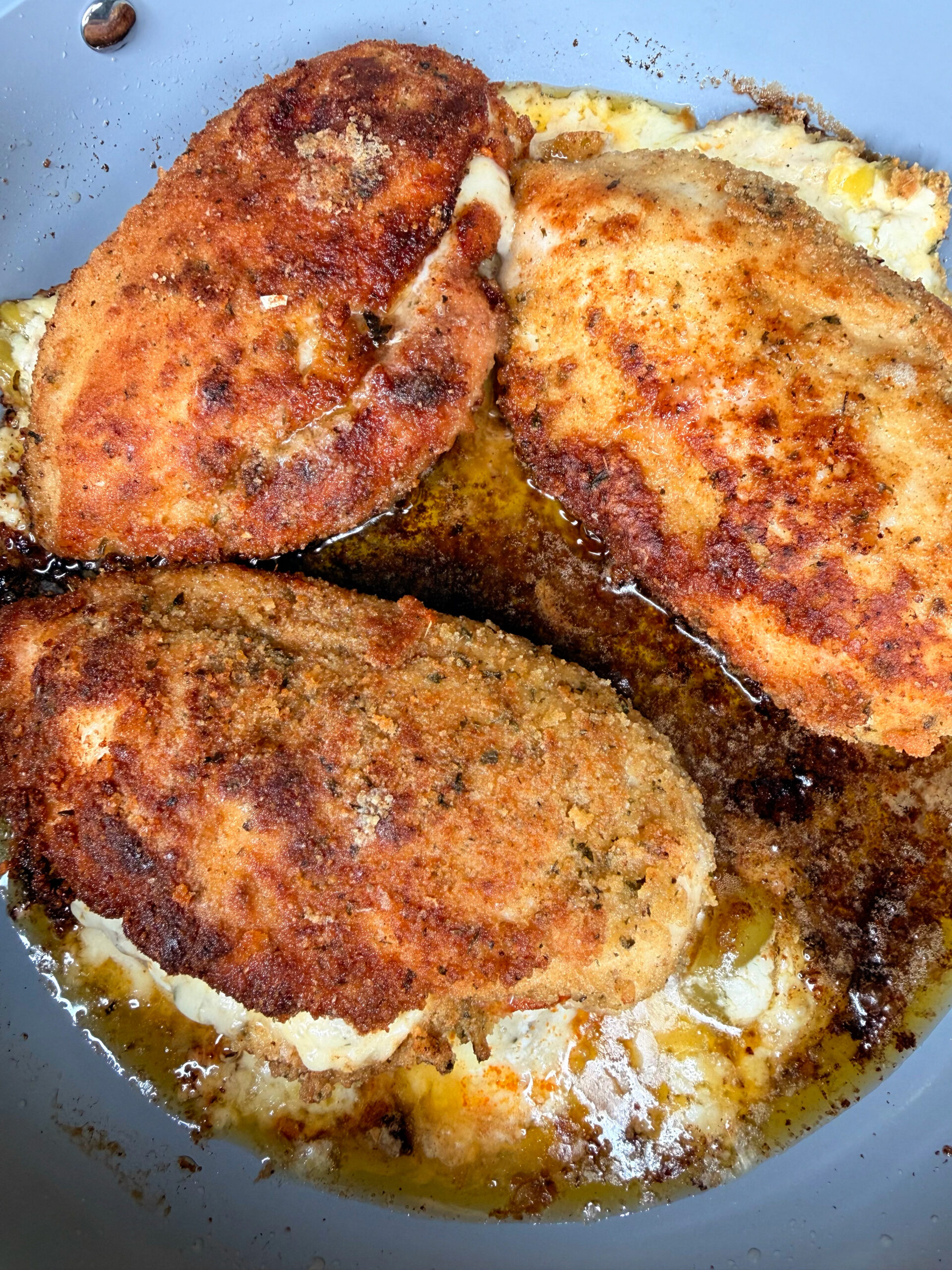 Three breaded and pan-fried chicken breasts are cooking in a skillet with sizzling oil and melted butter, creating a golden brown and crispy crust.