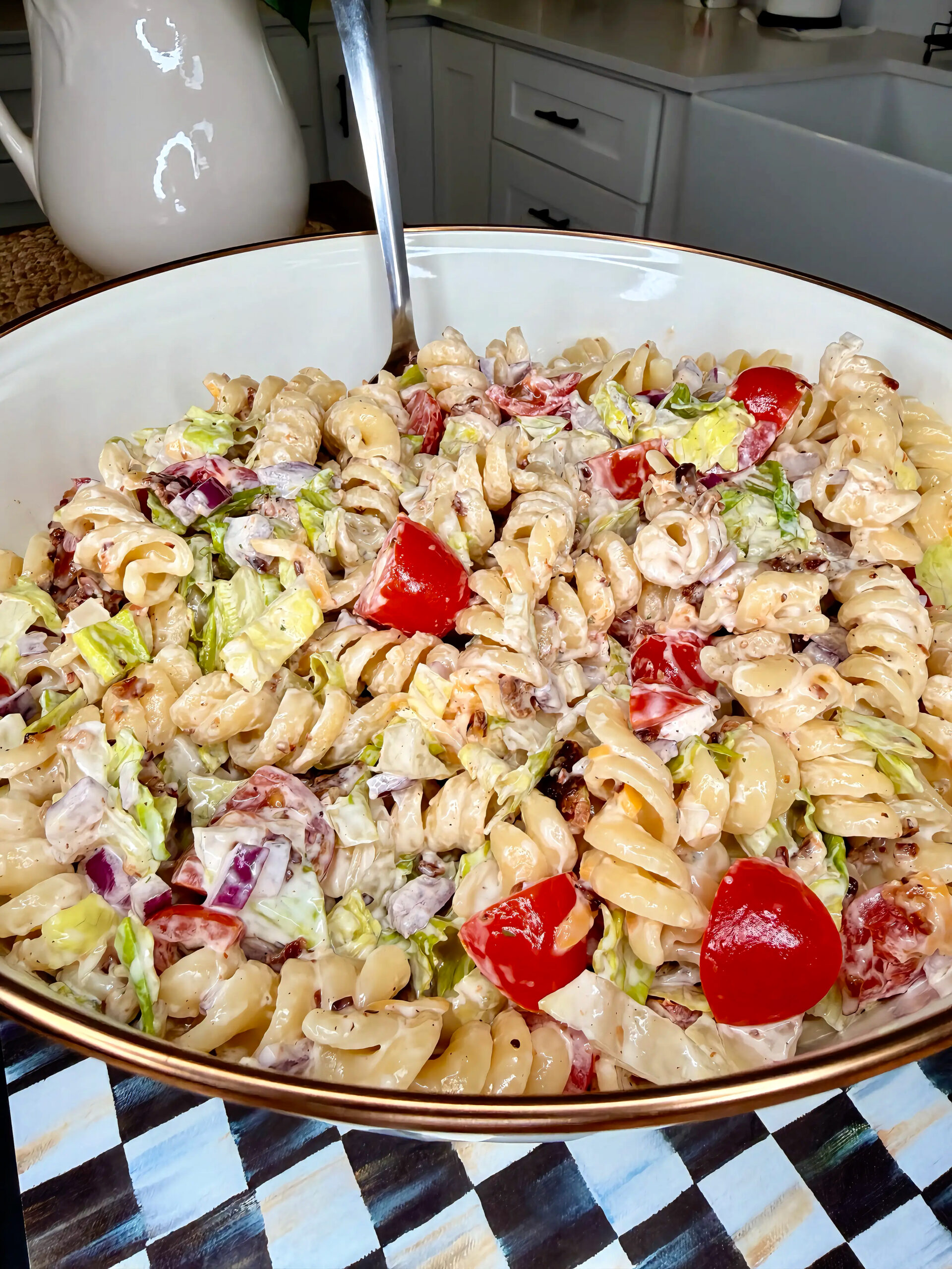 A large bowl of creamy pasta salad with rotini noodles, chopped lettuce, cherry tomatoes, red onions, and bacon on a kitchen counter. A serving spoon rests in the bowl.