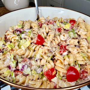 A large bowl filled with creamy rotini pasta salad, featuring chopped lettuce, grape tomatoes, red onion, and crispy bacon pieces, set on a checkered surface in a bright kitchen.