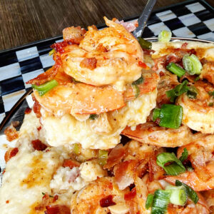 A close-up of shrimp and grits topped with bacon pieces and chopped green onions, with a fork lifting a bite. The dish is served on a patterned plate on a wooden table.