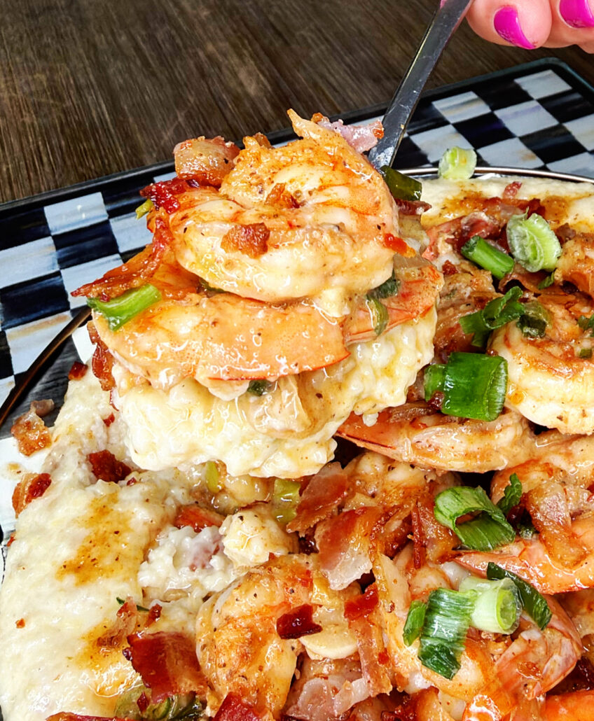 A close-up of shrimp and grits topped with bacon pieces and chopped green onions, with a fork lifting a bite. The dish is served on a patterned plate on a wooden table.