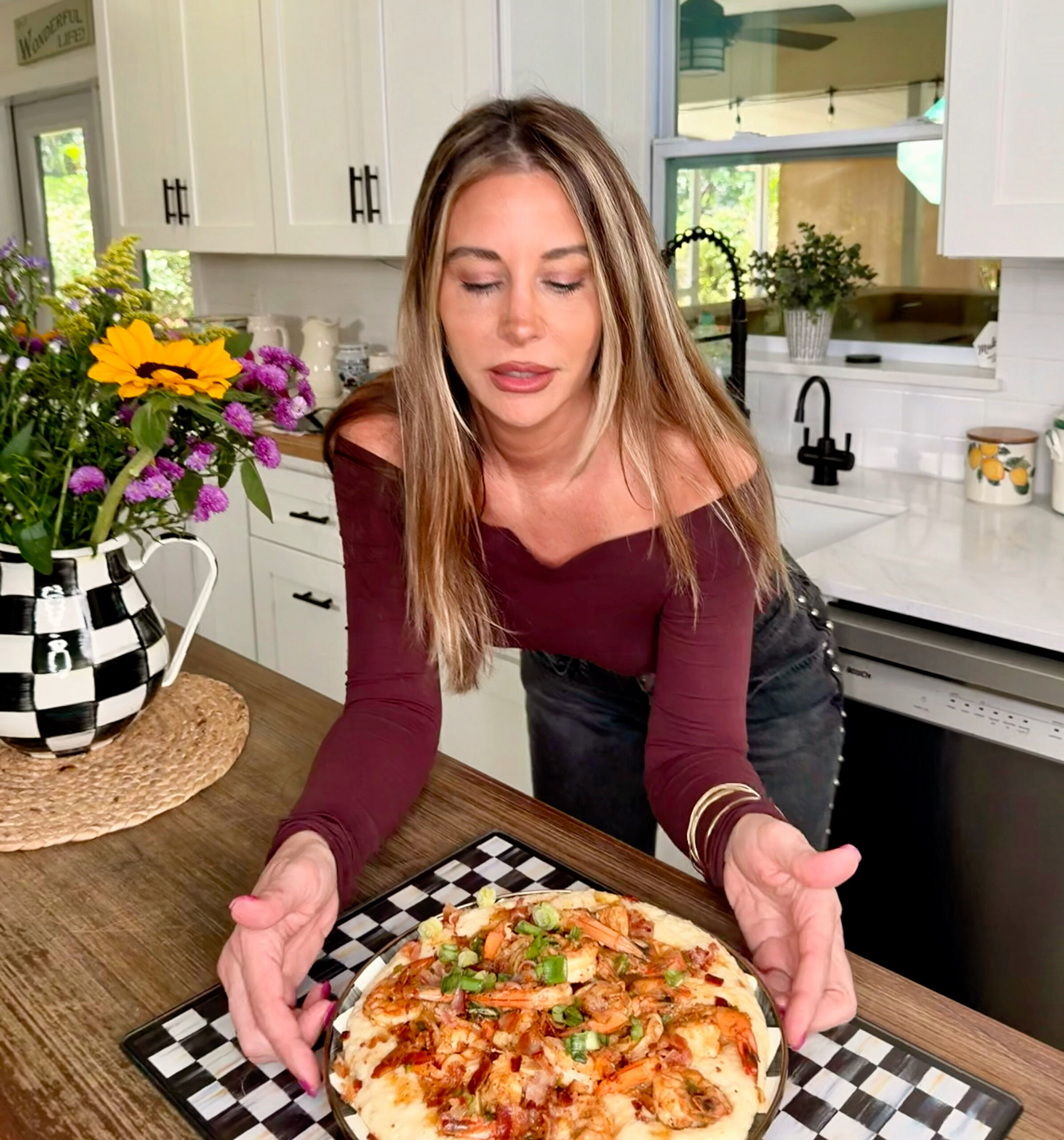 A woman with long brown hair in an off-shoulder maroon top presents a homemade pizza topped with vegetables in a bright, modern kitchen with flowers on the counter.