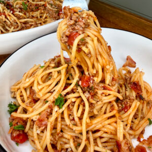 A bowl of spaghetti with meat sauce, garnished with chopped parsley and tomatoes. A fork is lifting a portion of the pasta from the bowl, with more pasta visible in a dish in the background.