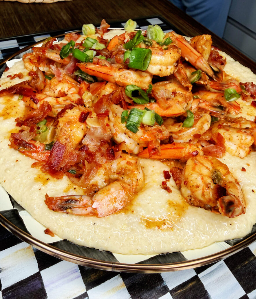 A plate of Creamy Southern Shrimp & Grits with Bacon Pan Sauce is topped with sautéed shrimp, crispy bacon pieces, and chopped green onions, all served on a decorative plate.
