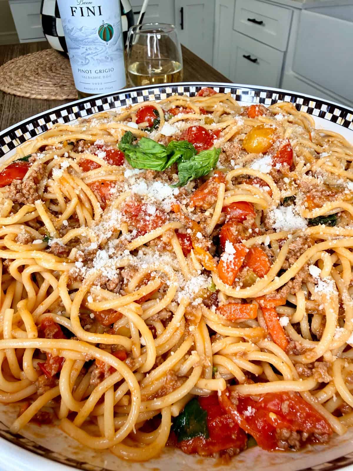 A close-up of a large bowl of spaghetti topped with ground meat, cherry tomatoes, basil, and grated cheese. A glass and a bottle of Pinot Grigio wine are in the background on a kitchen counter.