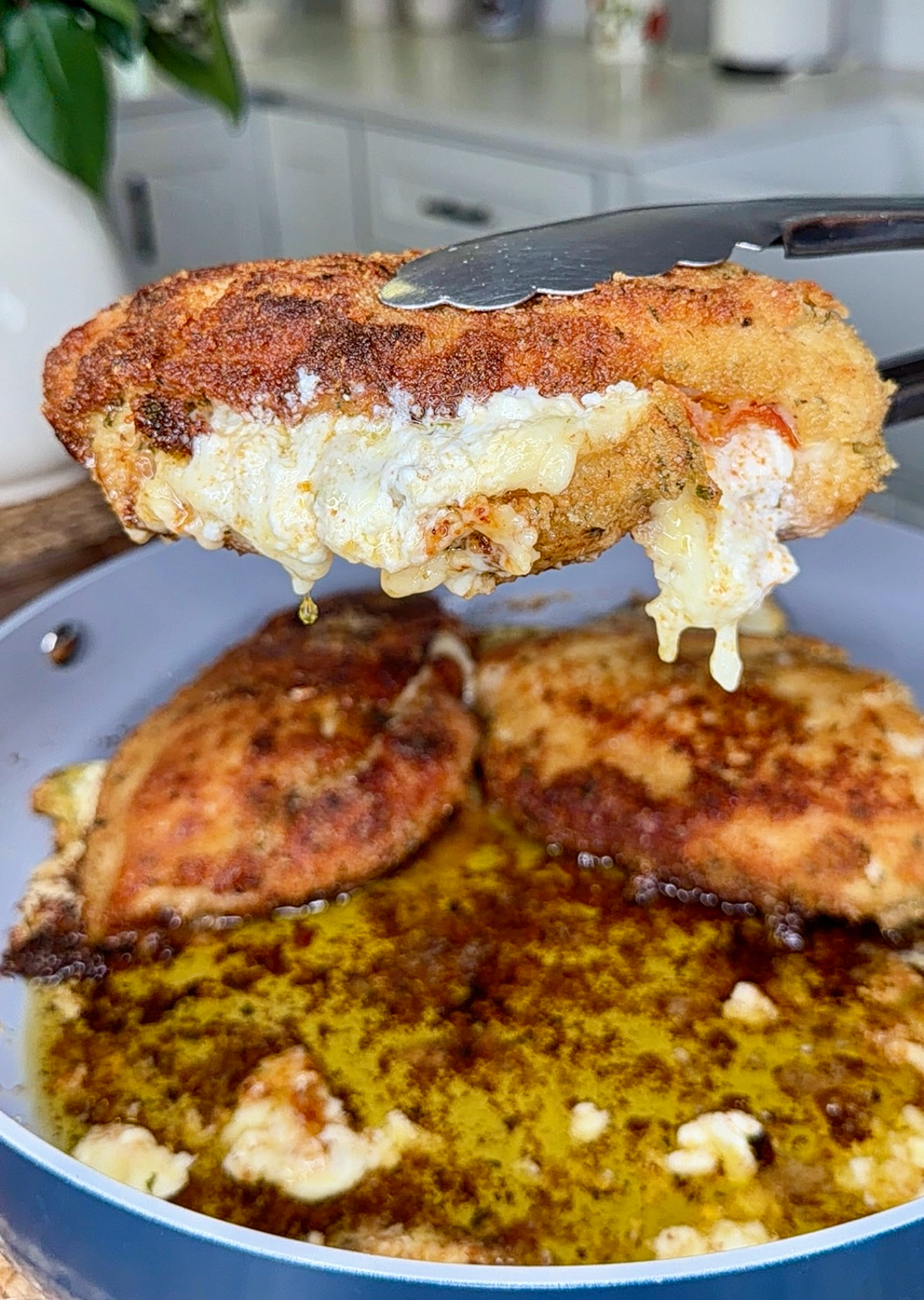 A close-up of tongs holding a breaded, fried chicken breast with melted cheese oozing out, above a pan with more pieces cooking in bubbling oil. The kitchen background is slightly blurred.