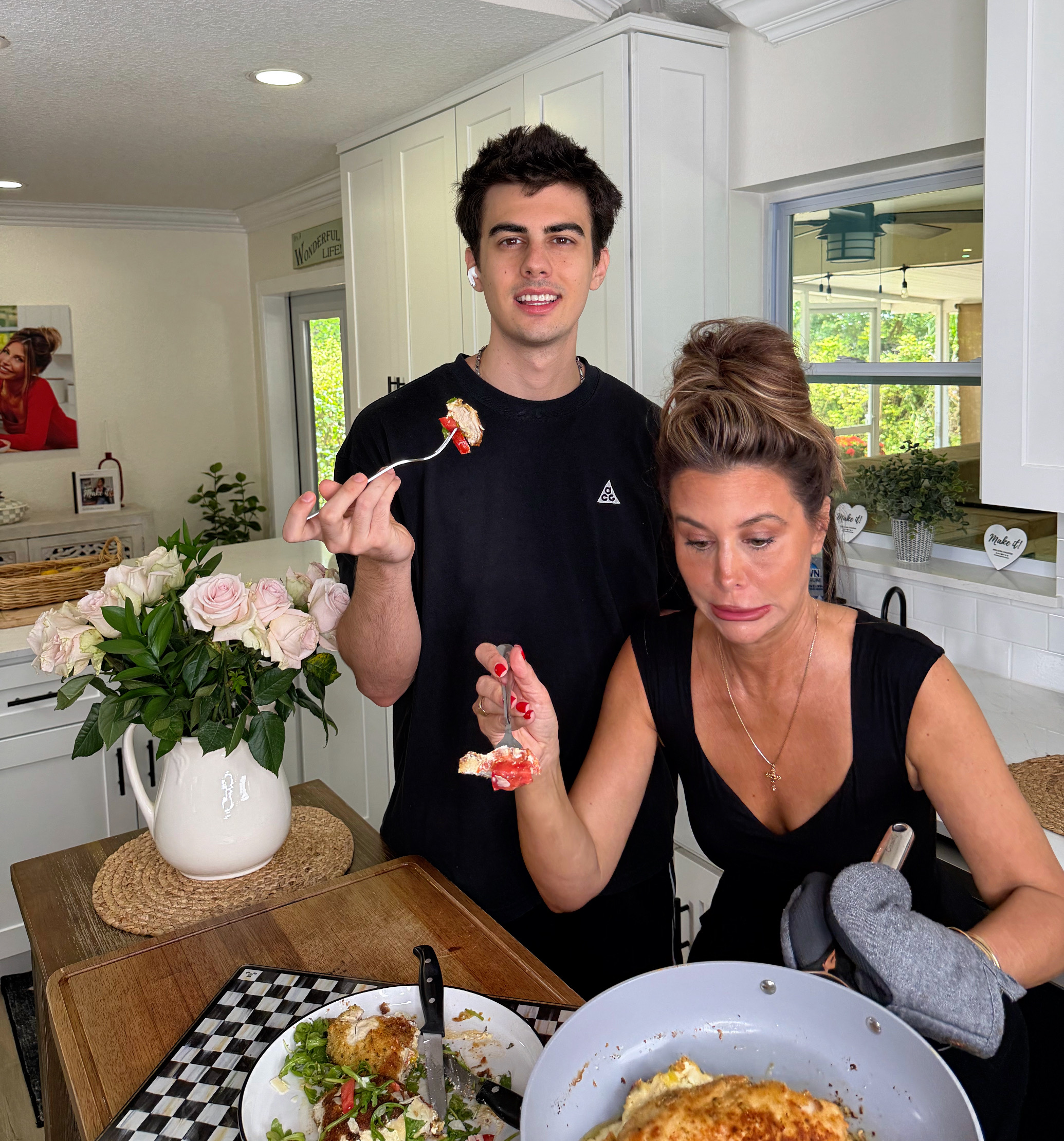 A young man and a woman stand in a bright kitchen, eating food. The woman holds a frying pan and wears an oven mitt. A vase of roses and a plate with food are on the table beside them. Both appear relaxed and casual.