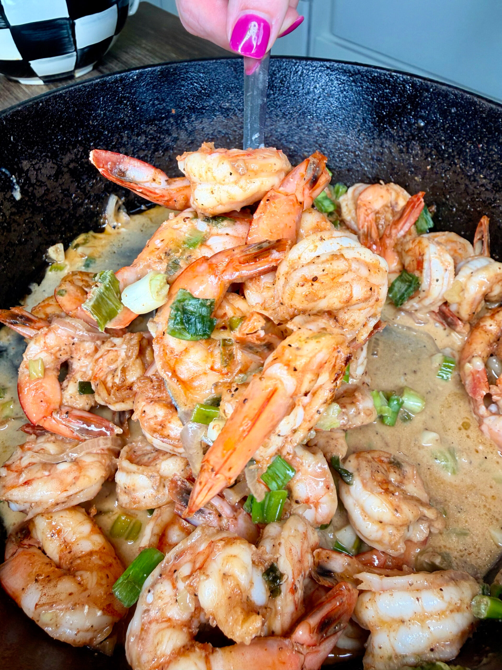 A close-up of a skillet filled with cooked shrimp, onions, and green onions in a creamy, seasoned sauce. A hand with pink nail polish holds a utensil lifting some shrimp from the pan.