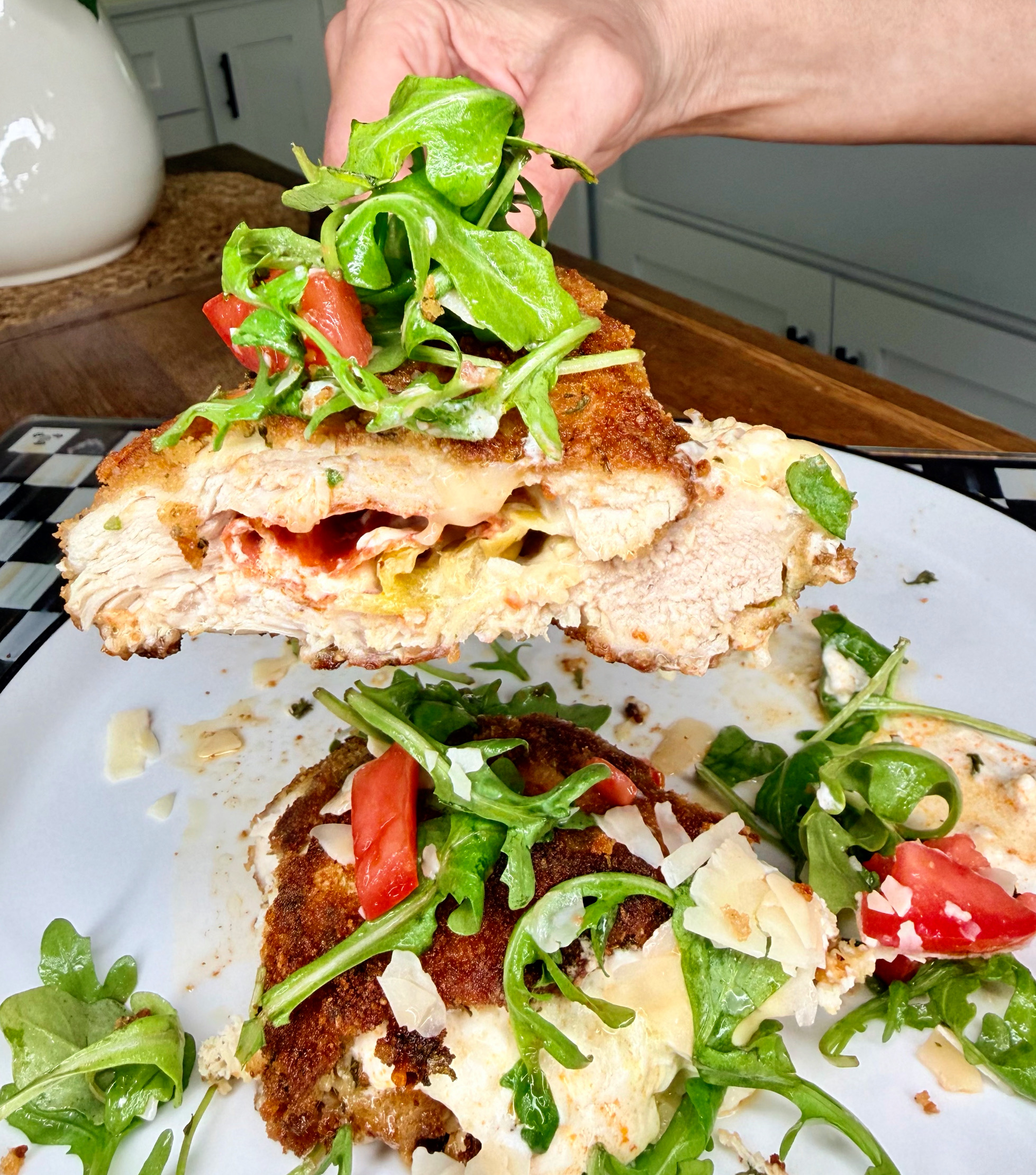A hand holds a breaded, stuffed chicken breast topped with arugula, tomato, and shaved cheese above a plate with more of the same dish on it, in a bright kitchen setting.