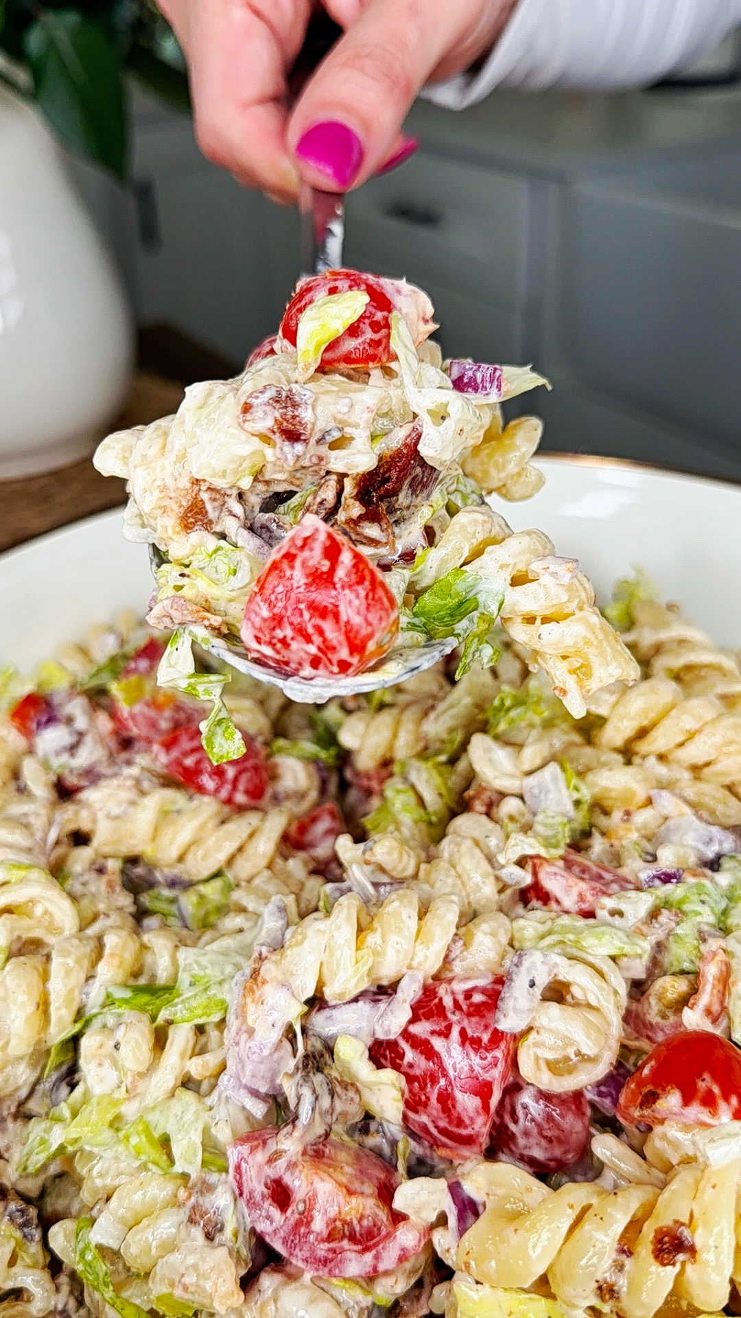 A close-up of a creamy pasta salad with rotini, cherry tomatoes, lettuce, red onions, and ground beef being lifted with a fork by a person with pink nail polish.