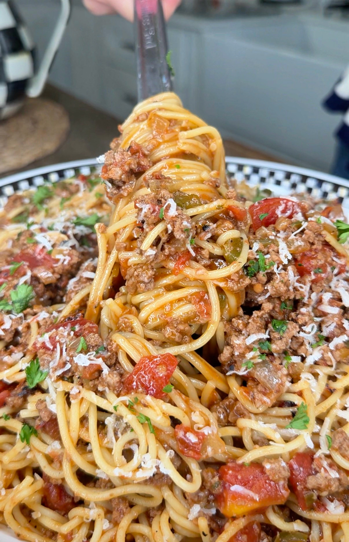 A close-up of spaghetti topped with meat sauce, diced tomatoes, grated cheese, and chopped parsley, being twirled around a fork above a bowl.