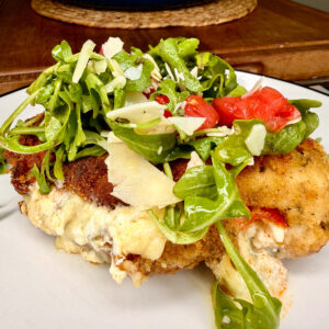 A breaded, cheese-stuffed chicken breast is topped with fresh arugula, sliced tomatoes, and shaved parmesan on a white plate, with a blue pot in the blurred background.