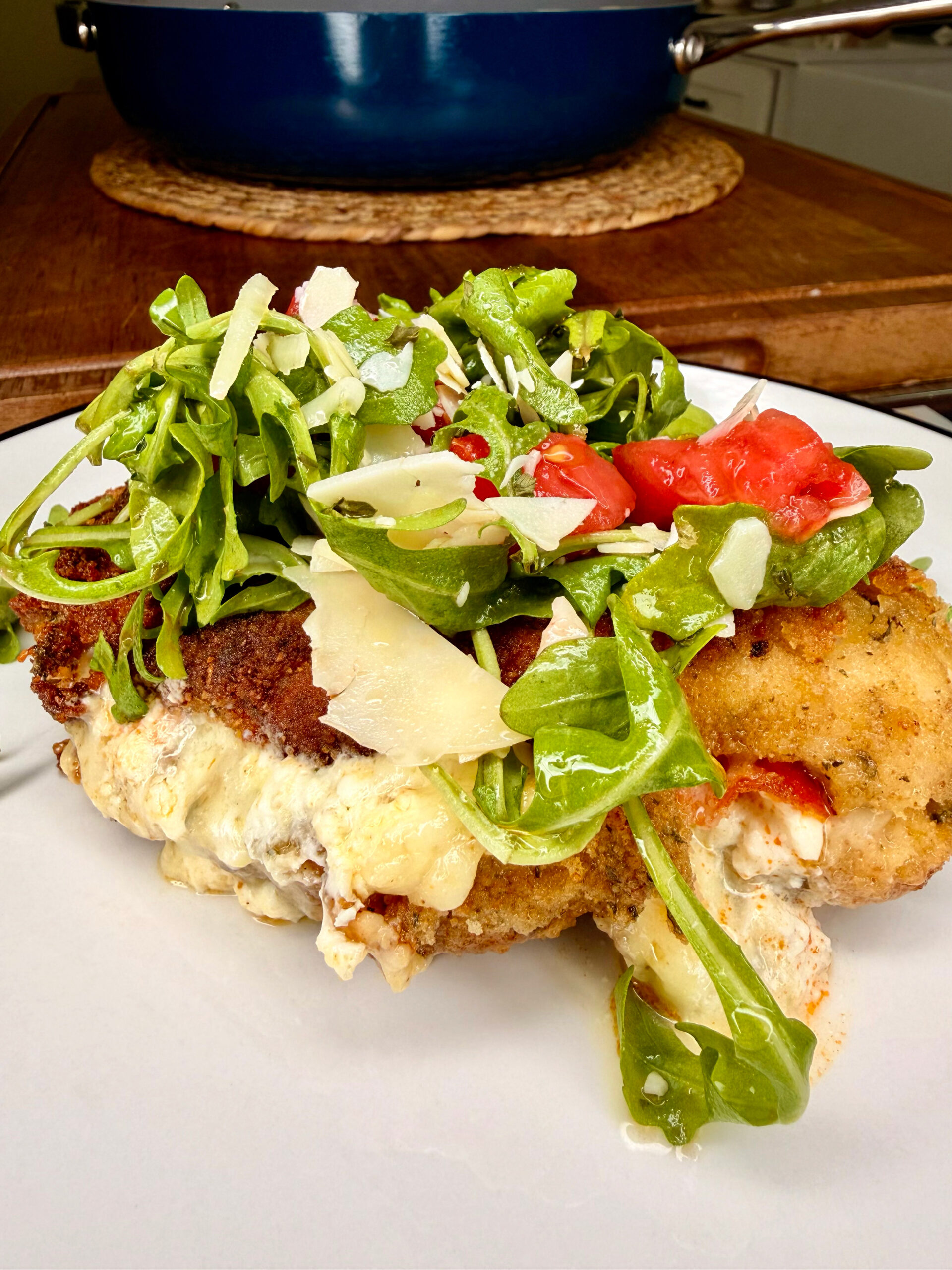 A breaded, cheese-stuffed chicken breast is topped with fresh arugula, sliced tomatoes, and shaved parmesan on a white plate, with a blue pot in the blurred background.