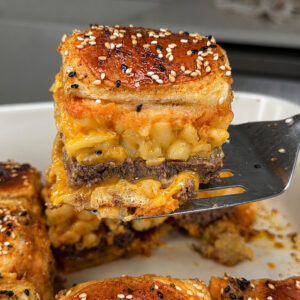 A spatula lifts a square sandwich from a baking dish, showing layers of ground beef and macaroni with cheese between golden, sesame-seed-topped buns. Other sandwiches remain in the dish on a kitchen counter.