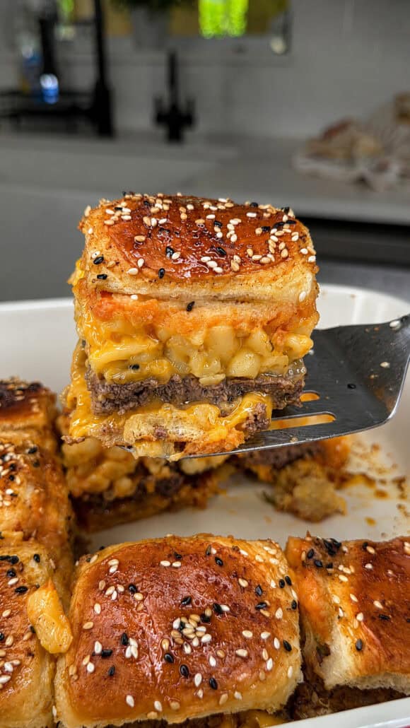 A spatula lifts a square sandwich from a baking dish, showing layers of ground beef and macaroni with cheese between golden, sesame-seed-topped buns. Other sandwiches remain in the dish on a kitchen counter.