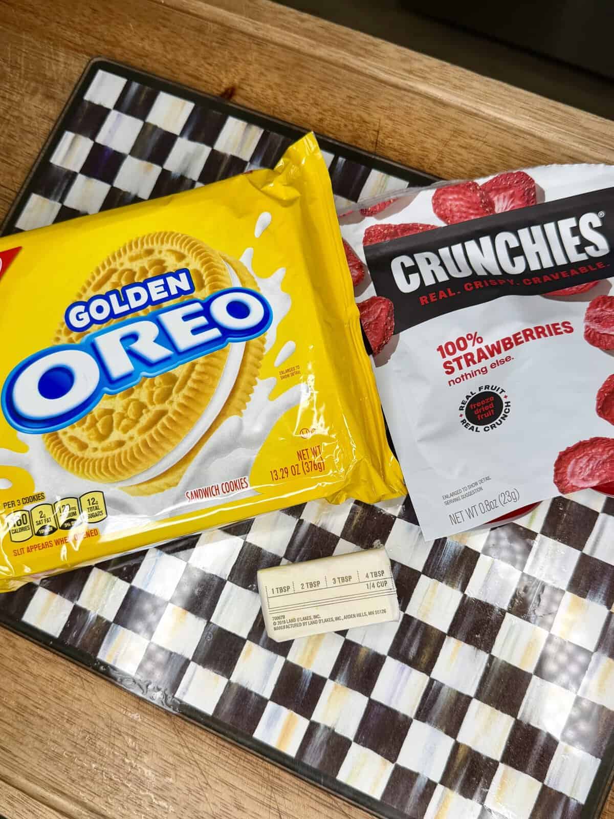 A package of Golden Oreo cookies, a bag of Crunchies freeze-dried strawberries, and a stick of butter sit on a black-and-white checkered cutting board on a wooden countertop.