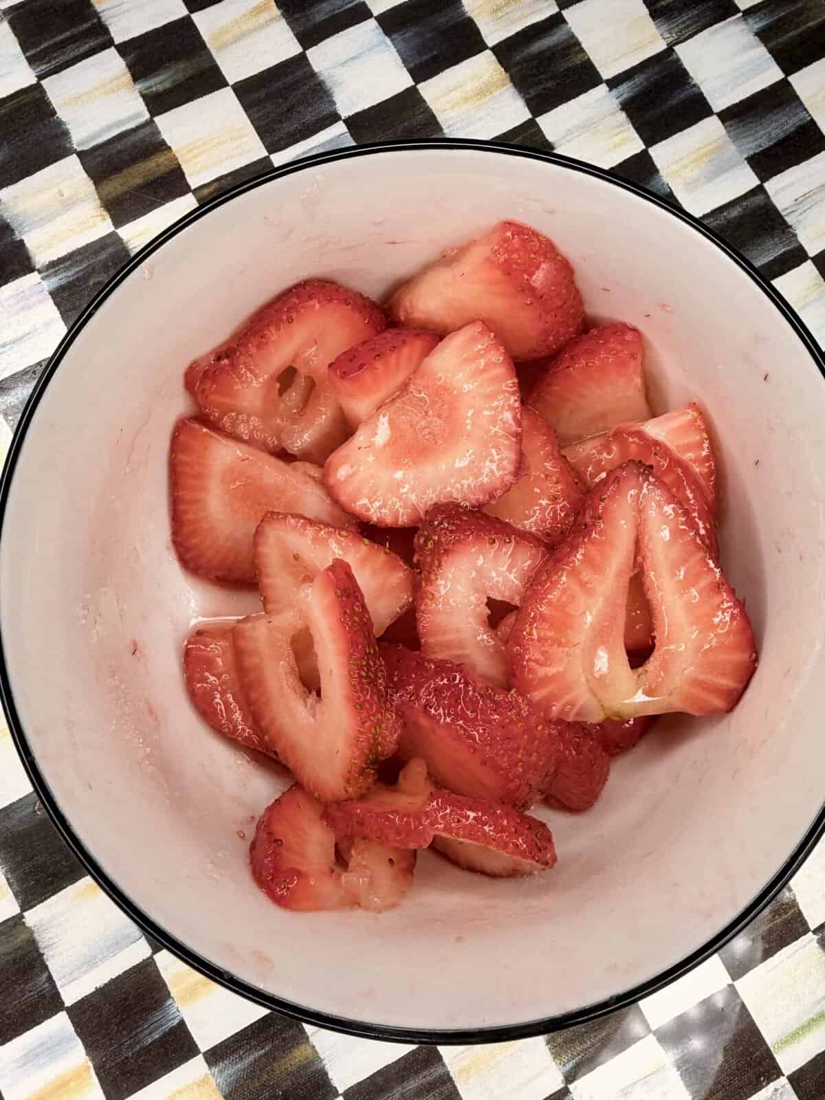 A white bowl filled with sliced strawberries sits on a checkered, multicolored surface. The strawberries are bright red and appear fresh and juicy.