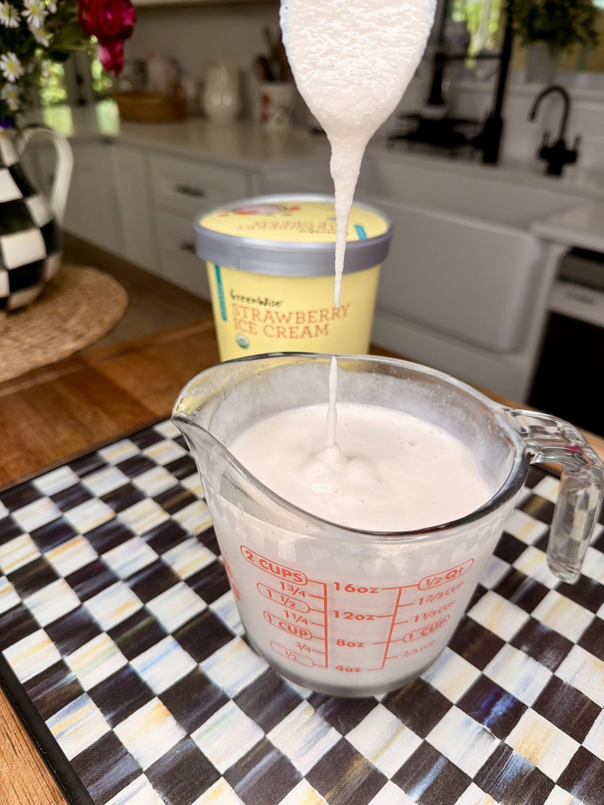 A thick, creamy mixture is being poured into a glass measuring cup on a checkered tray. In the background, there is a container of GreenWise Strawberry & Cream ice cream on a kitchen counter.